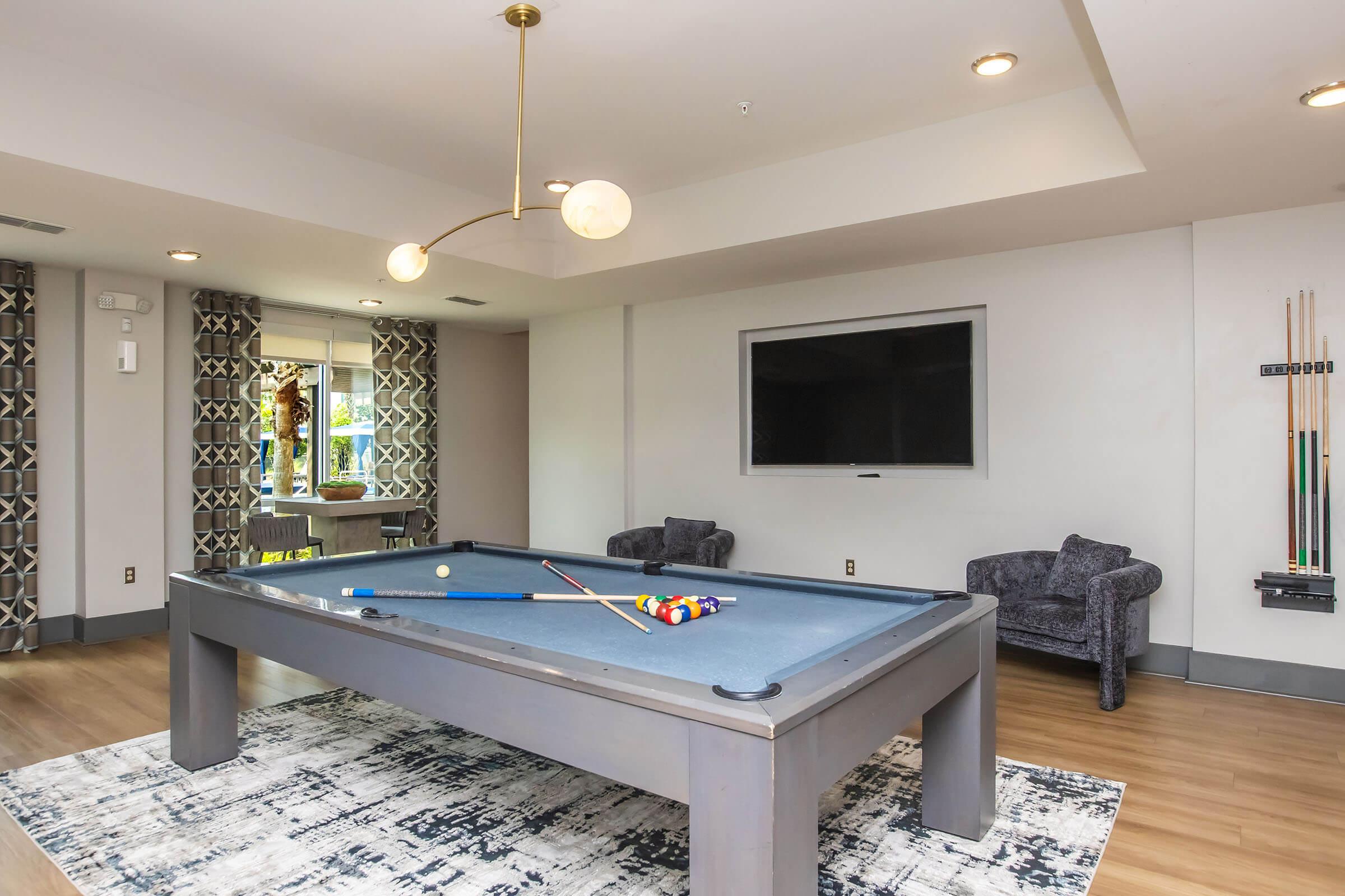 A cozy game room featuring a billiards table with colorful balls scattered on the table, two comfortable chairs in the corner, a large TV mounted on the wall, and modern decor including a stylish light fixture and patterned curtains. Hardwood flooring complements the relaxed atmosphere.