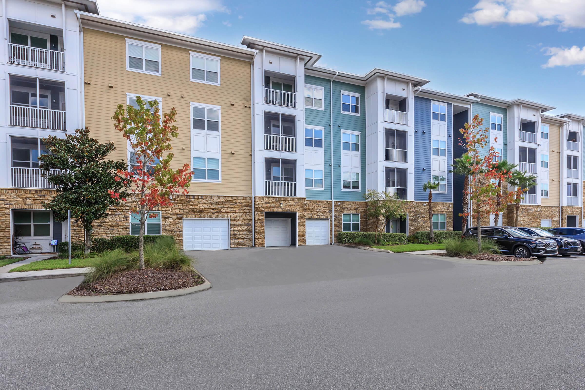 Modern residential apartment building with a mix of beige, blue, and white exteriors. The structure features multiple stories, balconies, and stone accents. Well-maintained landscaping includes small trees, and a parking area is visible in front, showcasing parked cars and a paved driveway.