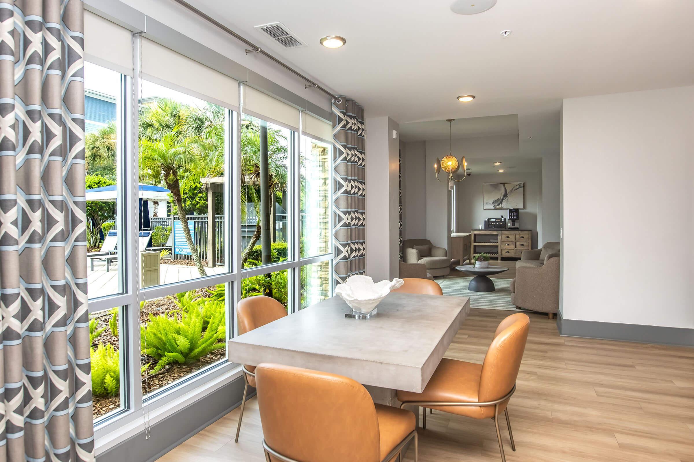 Modern dining area featuring a sleek gray table surrounded by four brown chairs. Large windows provide natural light and a view of a lush outdoor space with greenery. In the background, a cozy living area with light-colored furniture is visible, creating a stylish and inviting atmosphere.