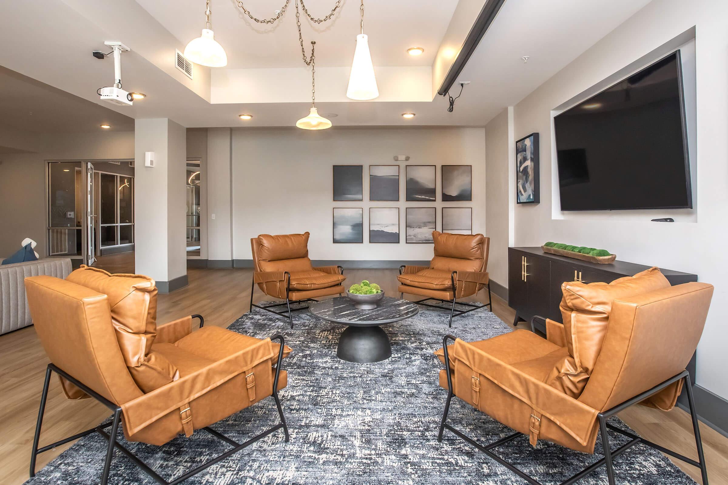 A modern lounge area featuring four brown leather chairs arranged around a black coffee table on a blue rug. A large television is mounted on the wall, and abstract framed artwork hangs above the seating. The space is brightly lit with pendant lighting and has a contemporary, inviting atmosphere.