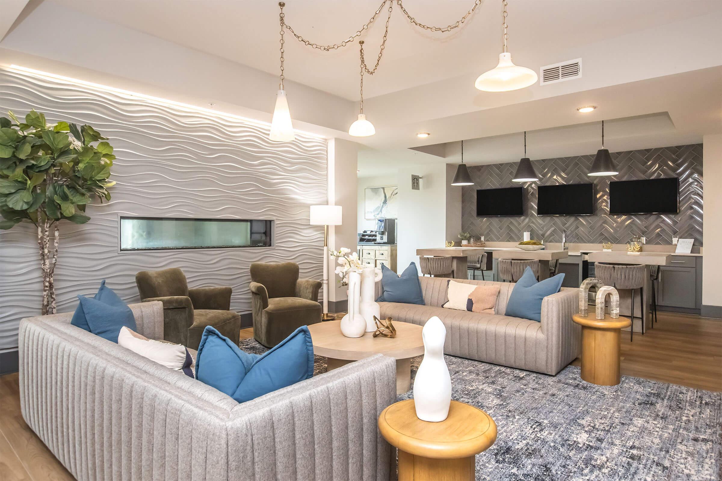 A modern living space featuring a textured wave-pattern wall, two green armchairs, a light gray sofa adorned with blue pillows, and a wooden coffee table. A decorative vase sits on the table. The area includes a dining space with two dark tabletops, four chairs, and two mounted screens, all complemented by pendant lighting.