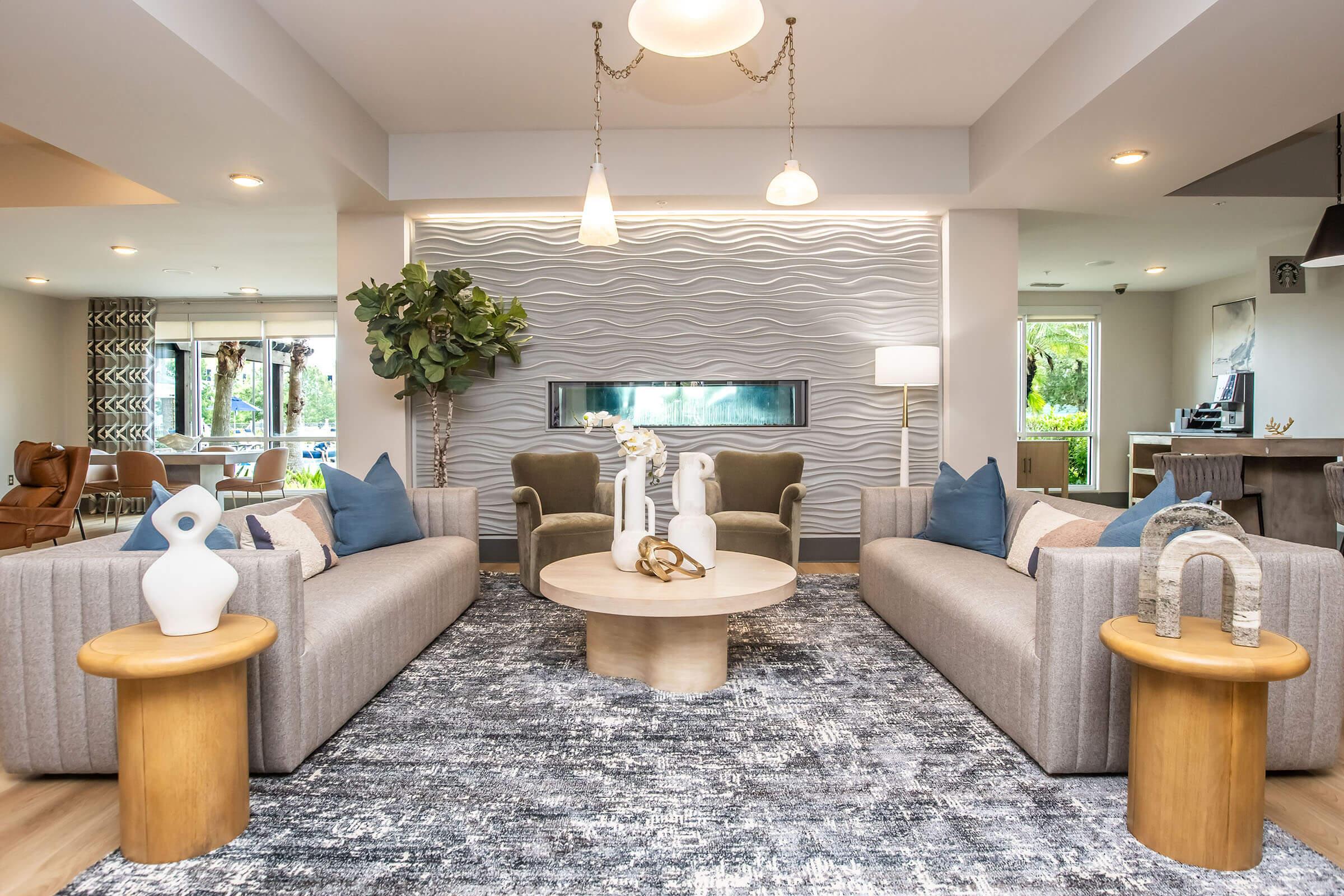 A modern living room featuring two gray sofas adorned with blue cushions, a round wooden coffee table in the center, and decorative vases on side tables. A textured wall serves as a backdrop, and there's a plant adding greenery, with large windows providing natural light.