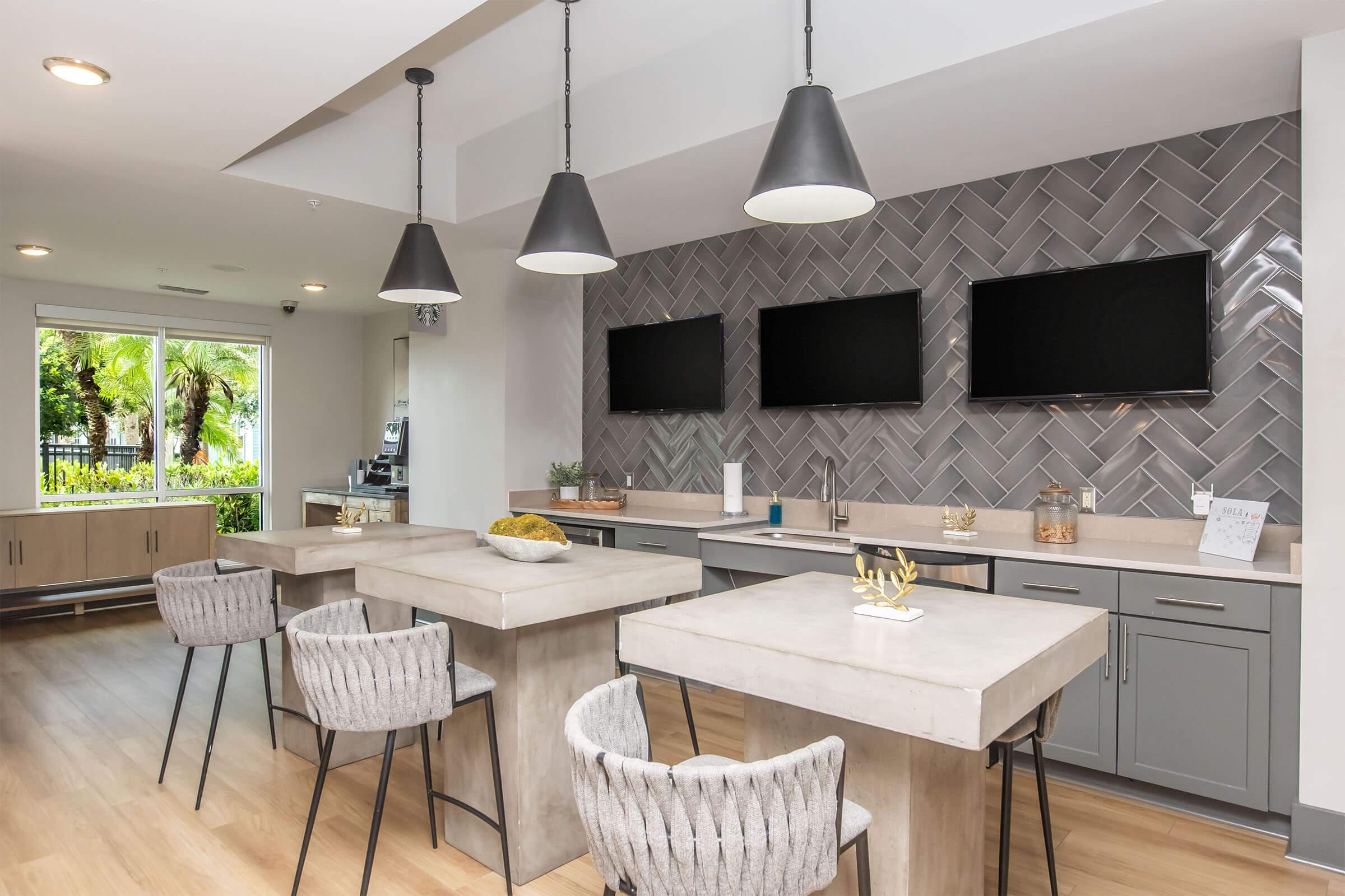 Modern kitchen area featuring a herringbone-patterned backsplash, two large islands with concrete tops, and stylish bar stools. Three flat-screen TVs mounted on the wall. Bright natural light coming from large windows overlooking greenery. Sleek cabinetry and decorative accents complete the contemporary design.