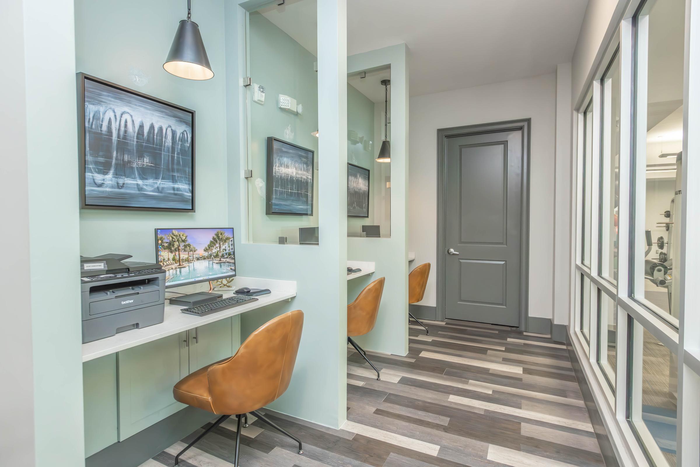 A modern coworking space featuring two workstations with sleek white desks and brown chairs. Each station has a computer monitor and printer. The room is well-lit with pendant lights and has decorative wall art. A glass wall allows natural light and visibility into the adjacent area.