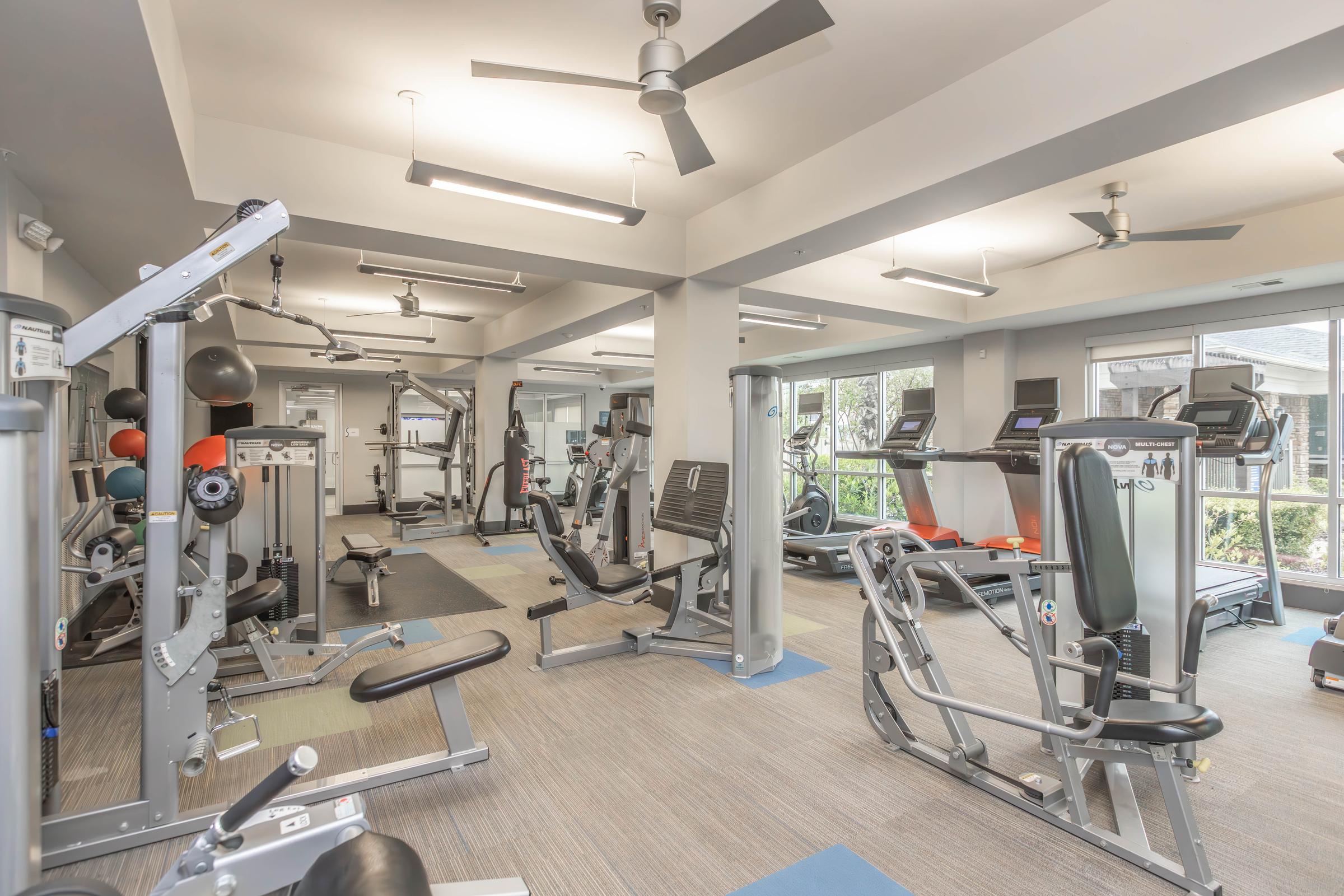 A modern gym interior featuring various exercise equipment including weight machines, treadmills, and stationary bikes. The space is well-lit with ceiling fans and large windows, providing a view of the outside. The flooring is carpeted in neutral tones, creating a welcoming atmosphere for workouts.