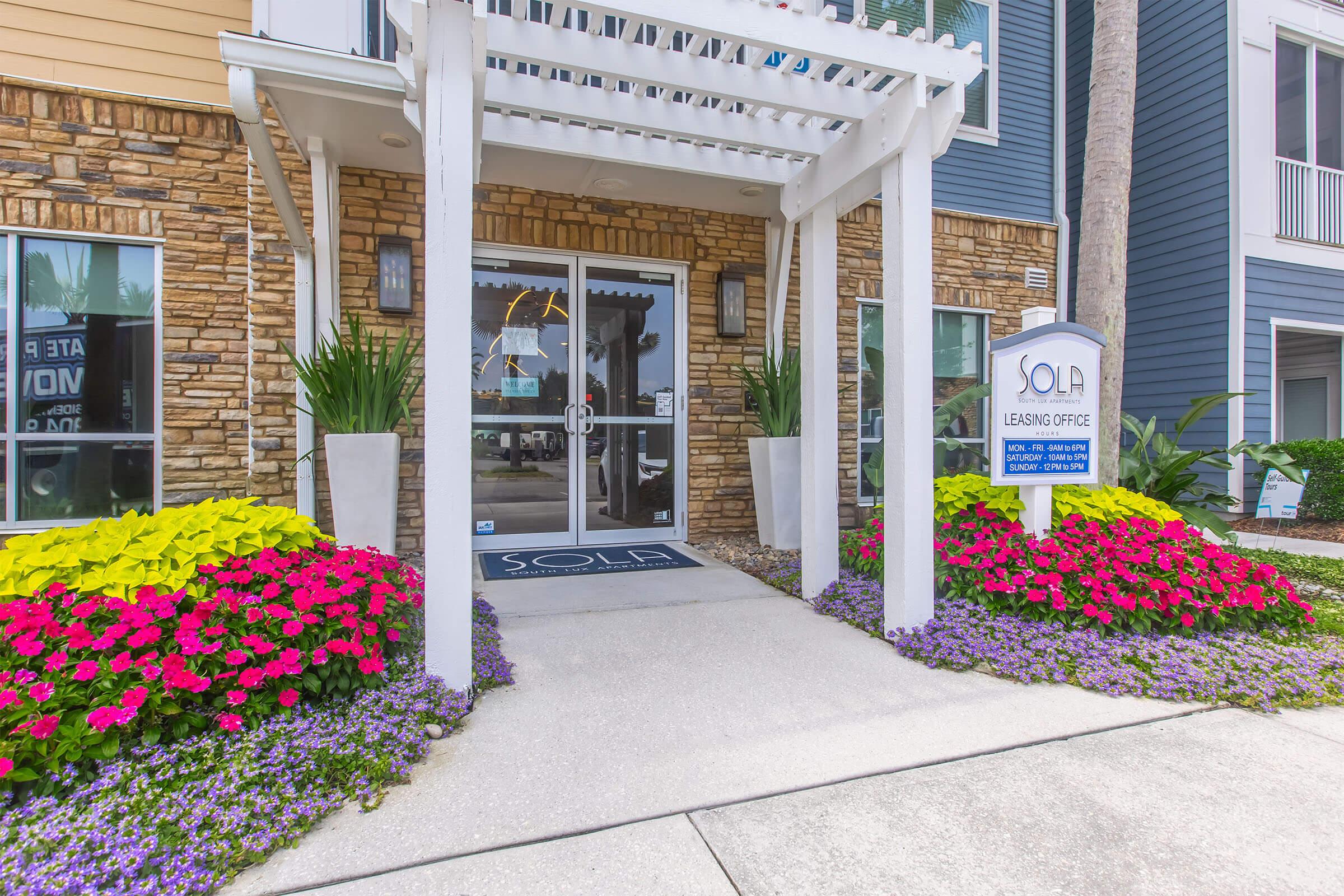Front entrance of a leasing office featuring a glass door, white wooden pergola, and vibrant flower beds with pink, yellow, and purple flowers. The office sign displays the name "SOLA" and additional leasing information. The building has a mix of stone and blue siding, creating a welcoming atmosphere.