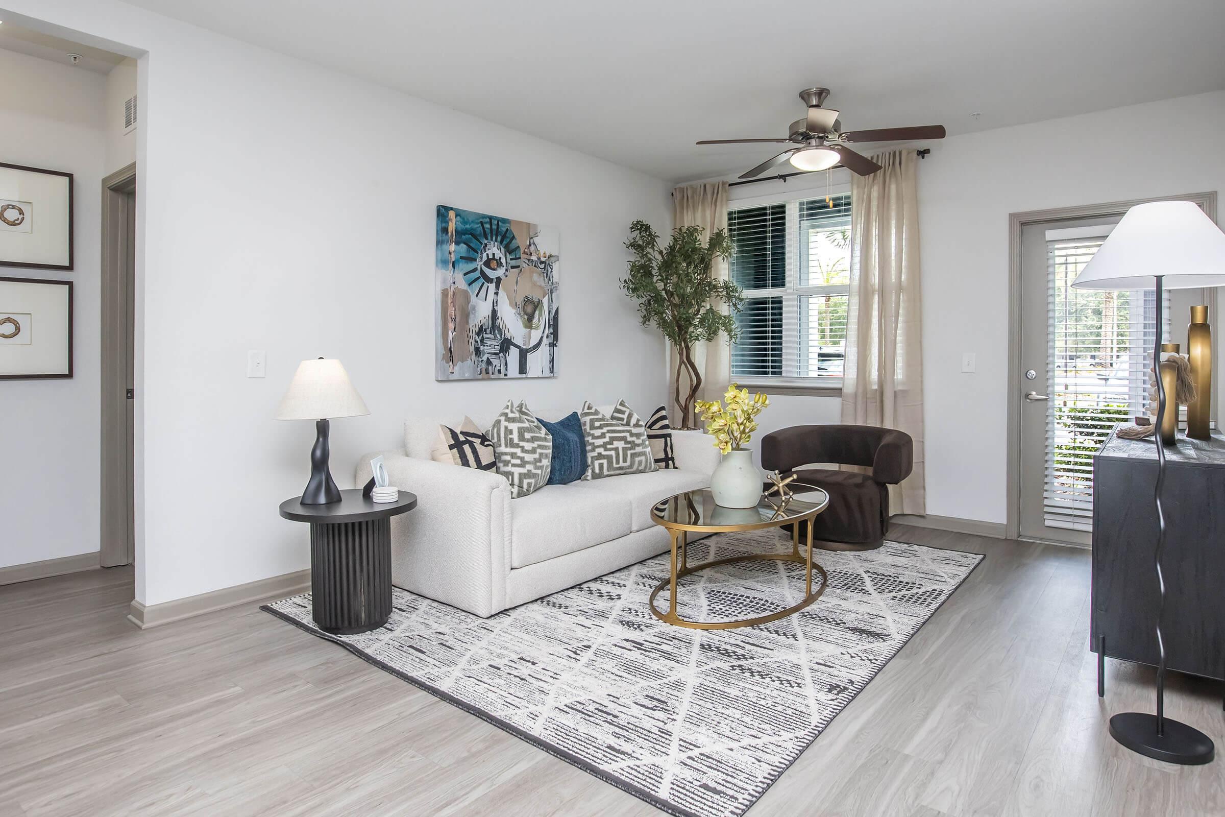 A bright and modern living room featuring a light-colored sofa adorned with patterned cushions, a round coffee table with a vase of yellow flowers, and a stylish armchair. Soft lighting from a table lamp complements the warm decor, while large windows allow natural light to fill the space.