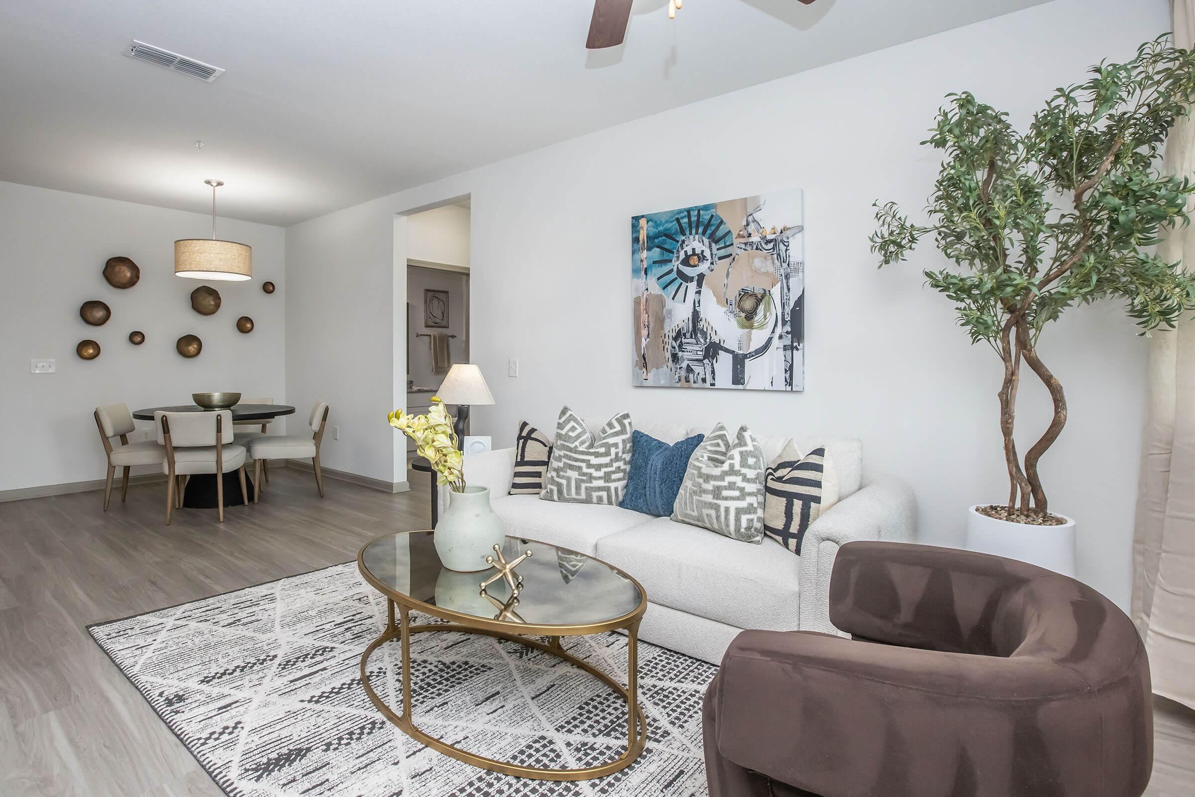 Living room featuring a light-colored sofa with patterned pillows, a round coffee table, and a decorative plant. In the background, there is a dining area with a round table and chairs, a modern chandelier, and wall decor. The walls are painted in neutral tones, creating a cozy atmosphere.