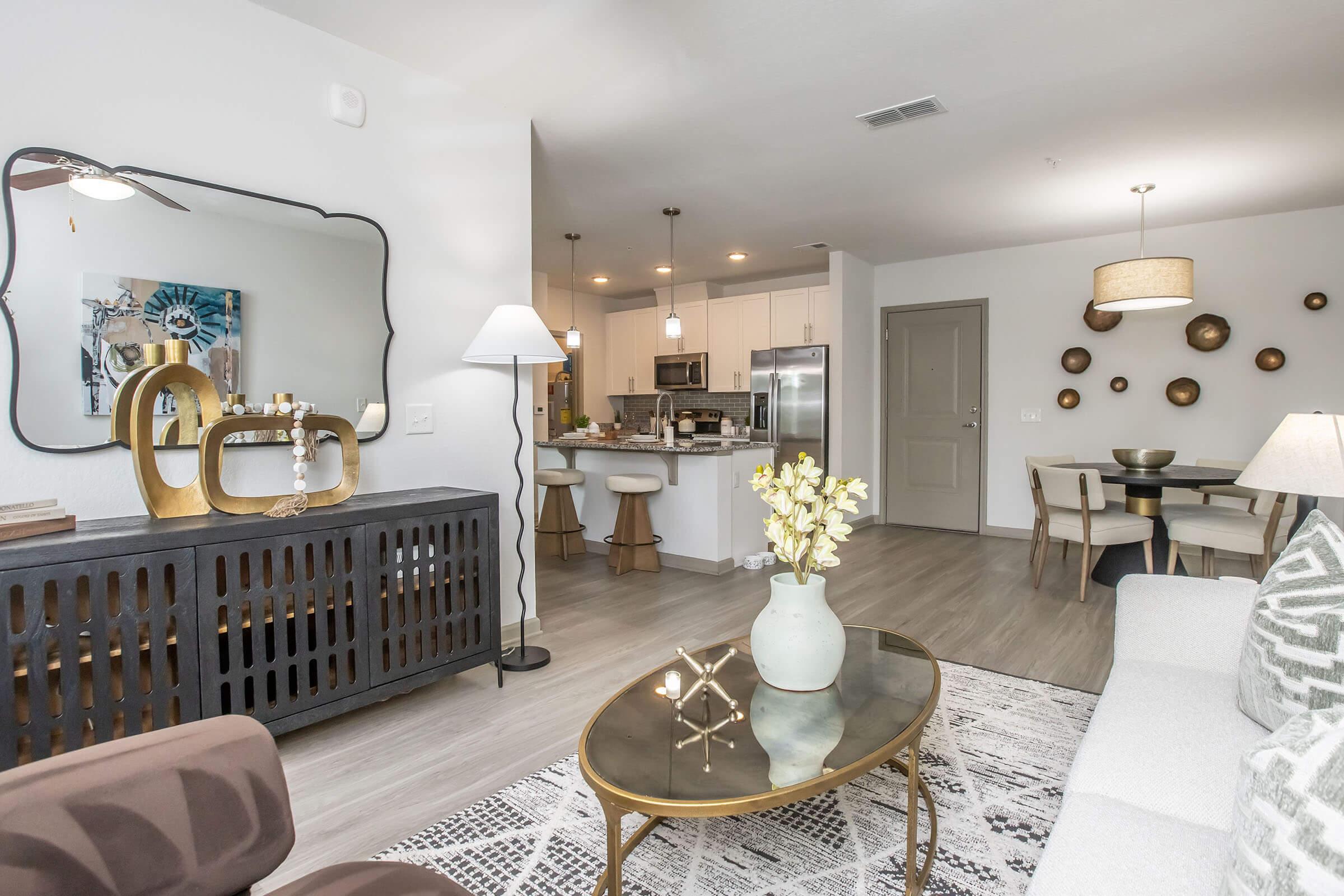 A modern living space featuring a stylish sofa, coffee table, and decorative elements. The room is well-lit with lamps, showcasing a dining area in the background. A mirror hangs on the wall, and there are plants and artwork adding to the contemporary decor.