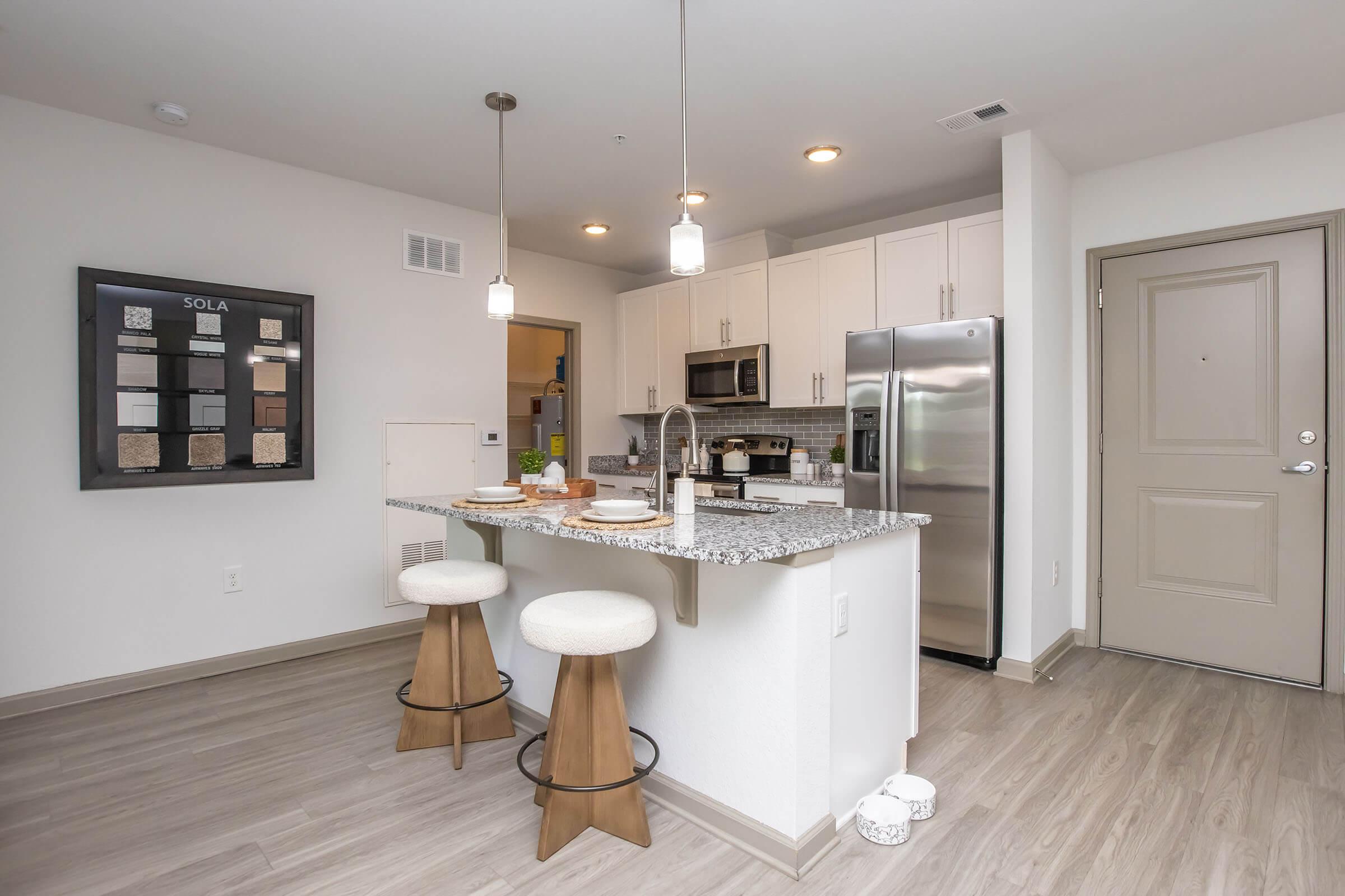 Modern kitchen with a granite island and two stools, featuring light cabinetry and stainless steel appliances. The space includes a framed board displaying various materials on the wall and a door leading out of the kitchen, with a light-colored wood floor throughout.