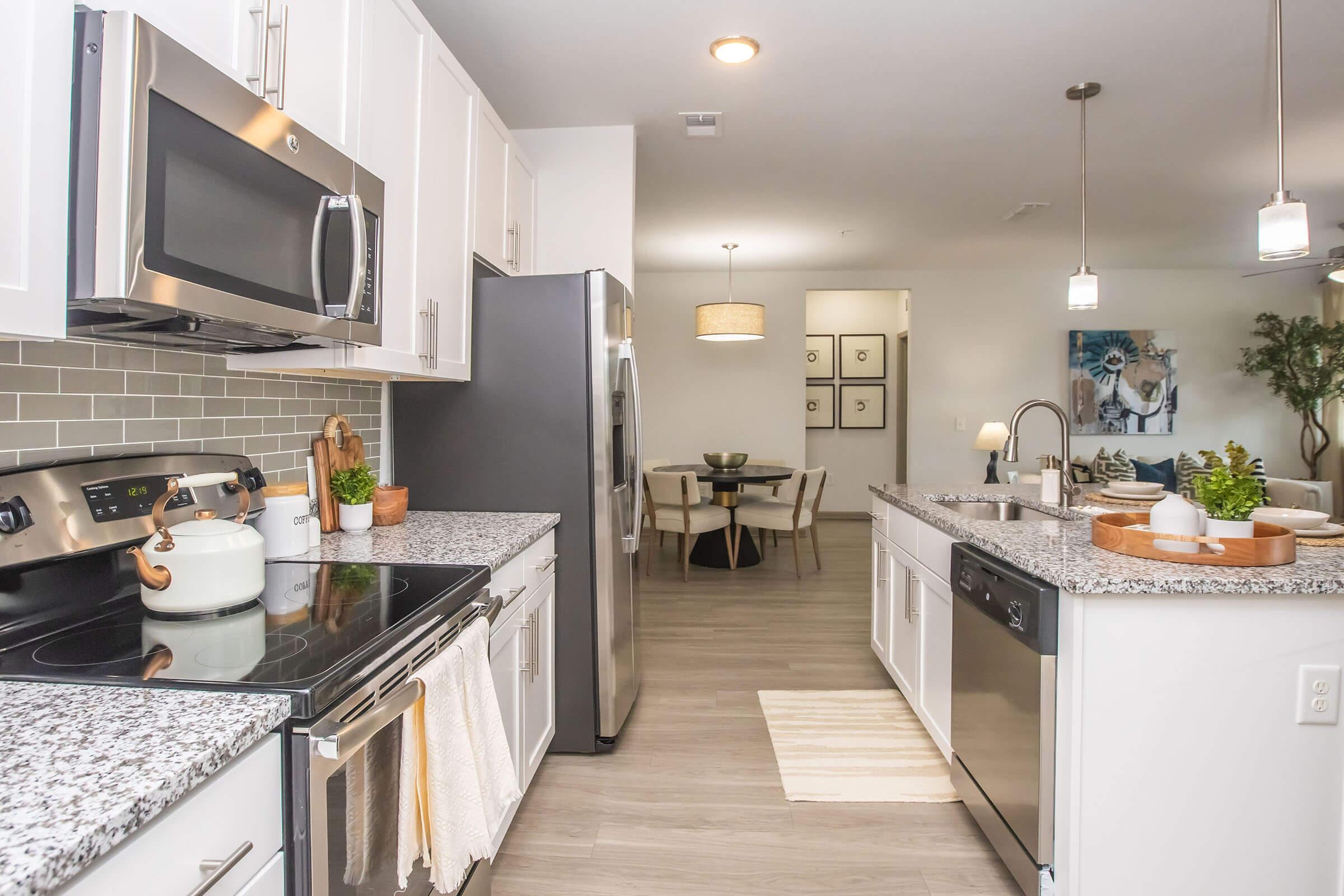 Modern kitchen with stainless steel appliances, including a microwave and dishwasher, paired with white cabinets and a gray granite countertop. There's a decorative plant on the counter, and a dining area with a light fixture in the background. The flooring is light wood, creating a bright, inviting atmosphere.
