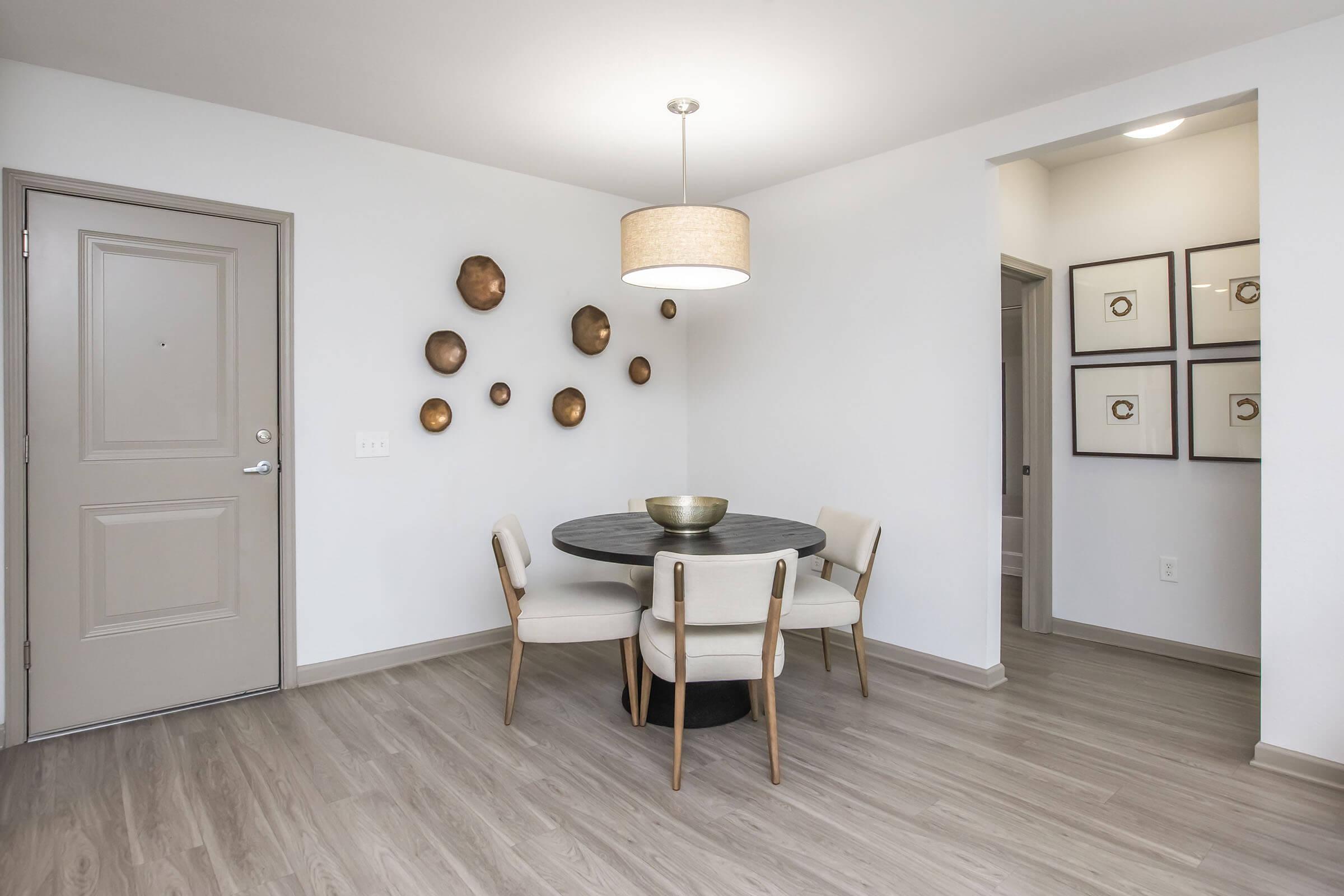 A modern dining area featuring a round black table with two beige chairs, a decorative bowl in the center, and a stylish light fixture above. The walls are adorned with circular wall art, and a closed door leads to another room, all set against a backdrop of light-colored walls and wooden flooring.