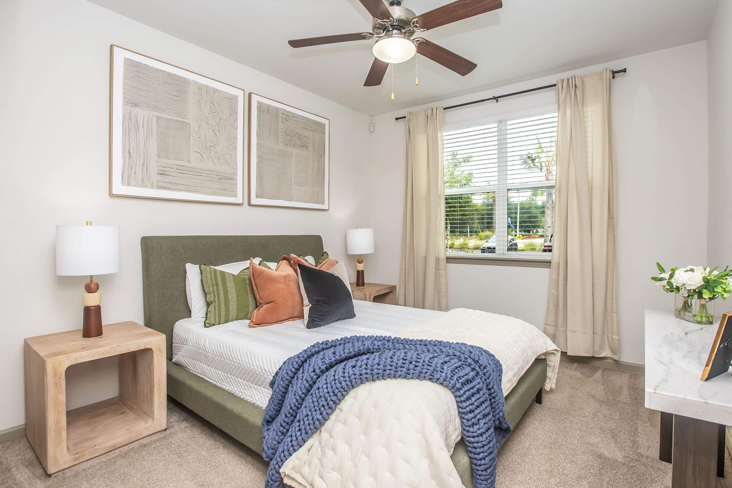 A cozy bedroom featuring a green upholstered bed with decorative pillows, a blue throw blanket, and soft lighting from bedside lamps. The walls are adorned with framed artwork, and there are sheer curtains over a window that lets in natural light. A small flower vase sits on a marble-topped nightstand.