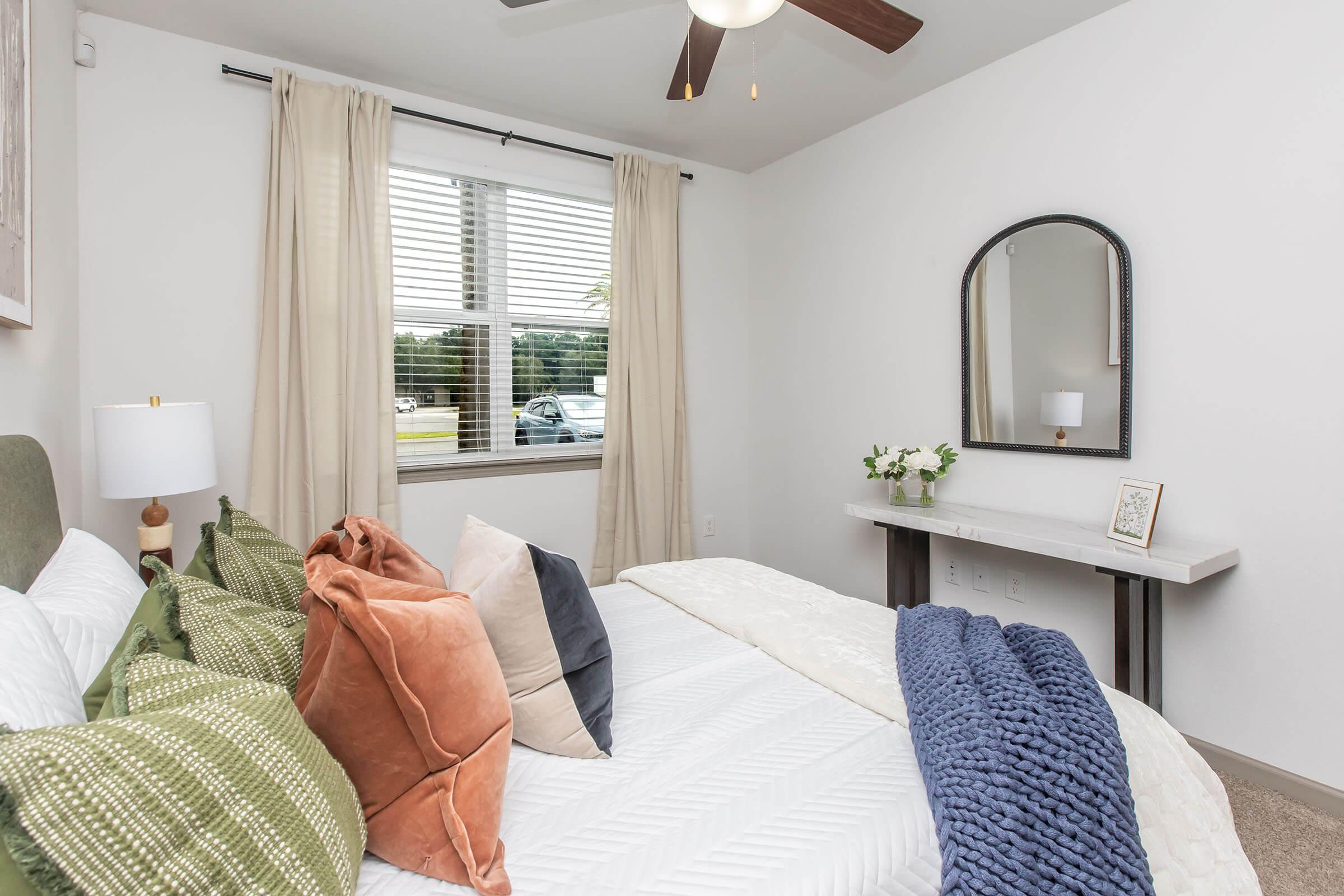 A cozy bedroom featuring a bed with decorative pillows in various colors, a stylish gray throw blanket, and a minimalist white bedside table. Natural light enters through a window with blinds, and a round mirror hangs above the table. Soft curtains enhance the inviting atmosphere of the space.