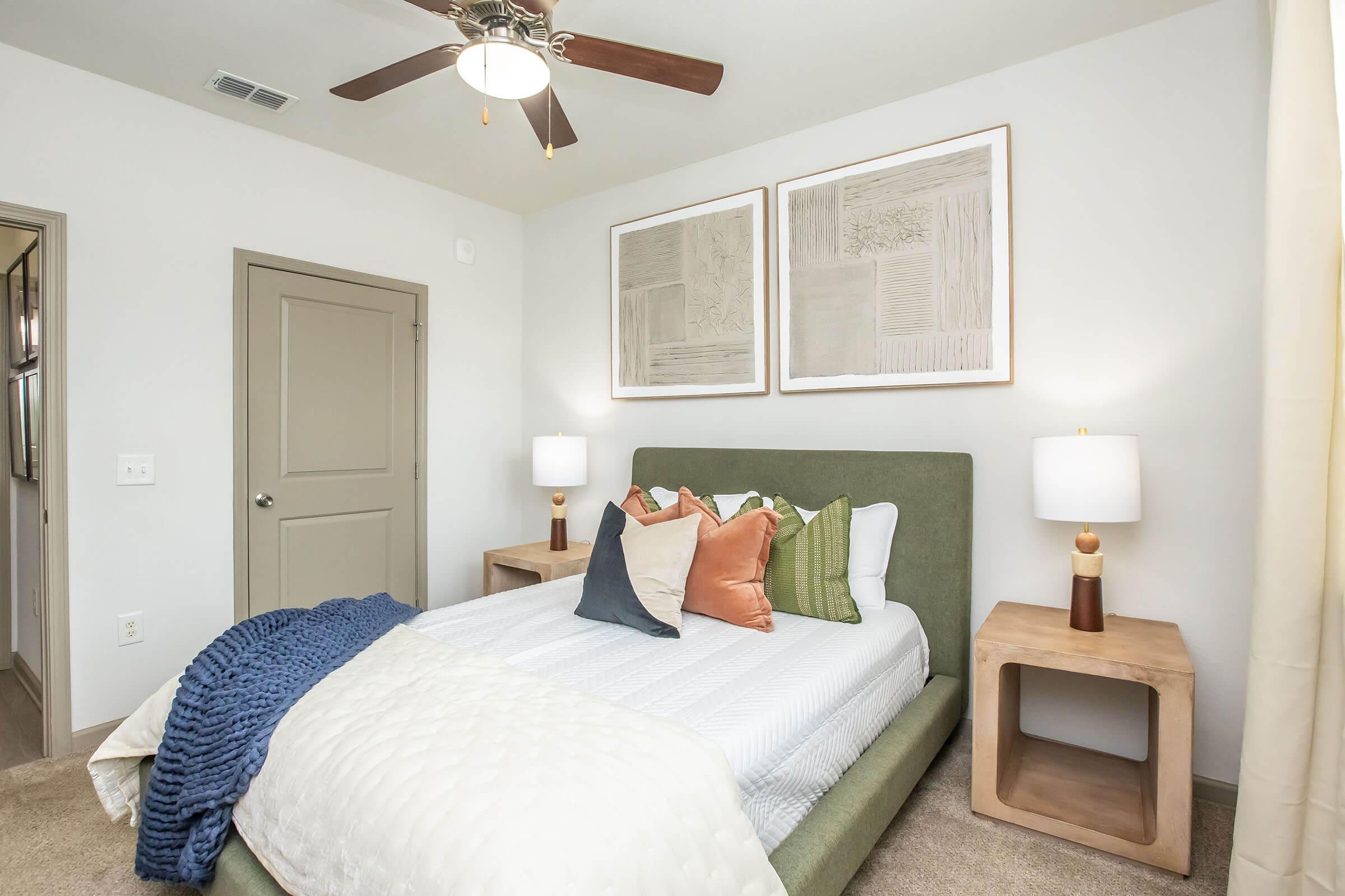 A cozy bedroom featuring a green upholstered bed with decorative pillows, two wooden bedside tables with lamps, and framed wall art. Soft lighting and neutral tones create a warm and inviting atmosphere, complemented by a ceiling fan and a plush throw blanket.