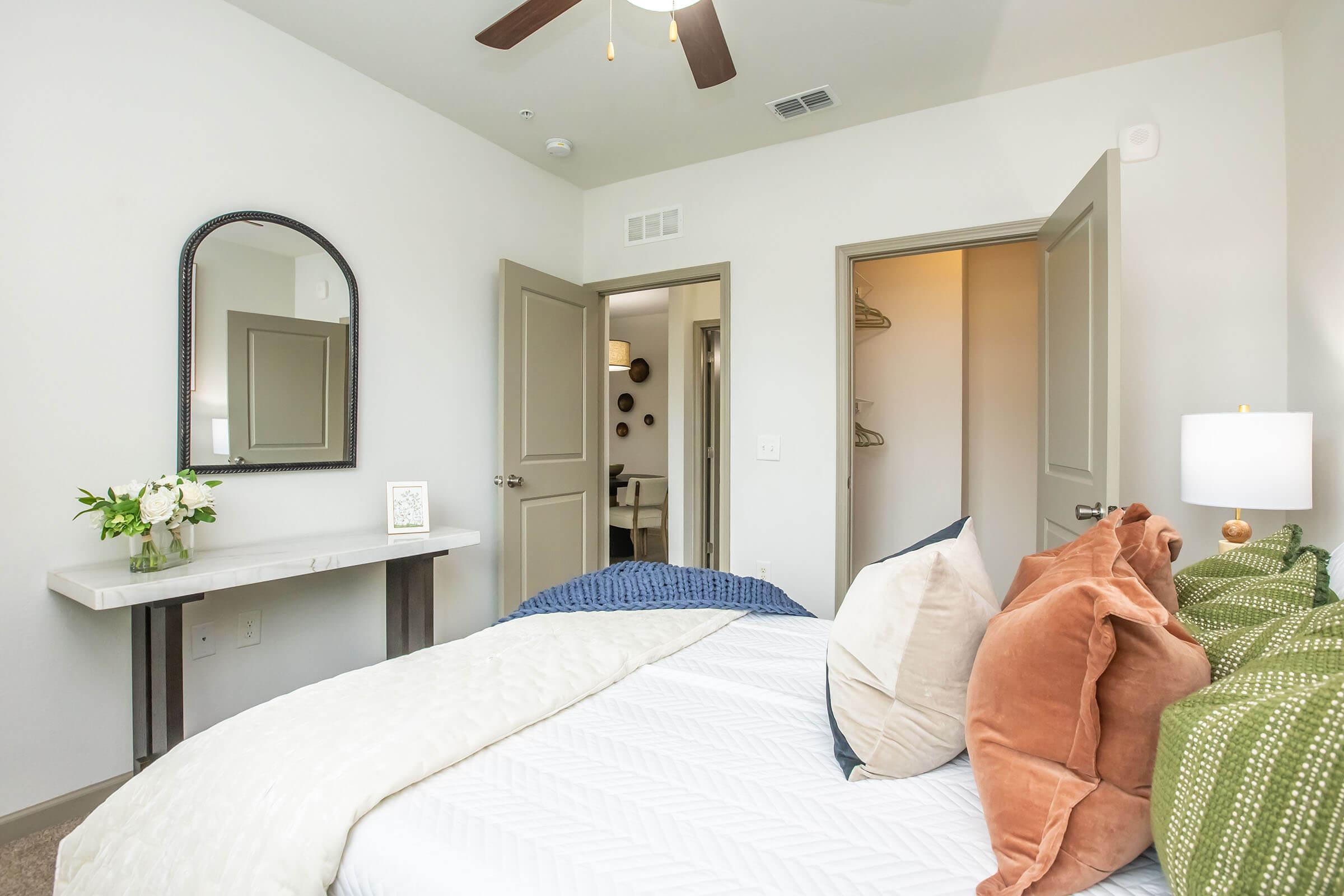 A well-decorated bedroom featuring a bed with decorative pillows and a cozy blanket. A small table with a floral arrangement and a mirror is positioned beside the bed. The room has light-colored walls, a ceiling fan, and a view of a closet through an open door. Soft, neutral tones create a welcoming atmosphere.