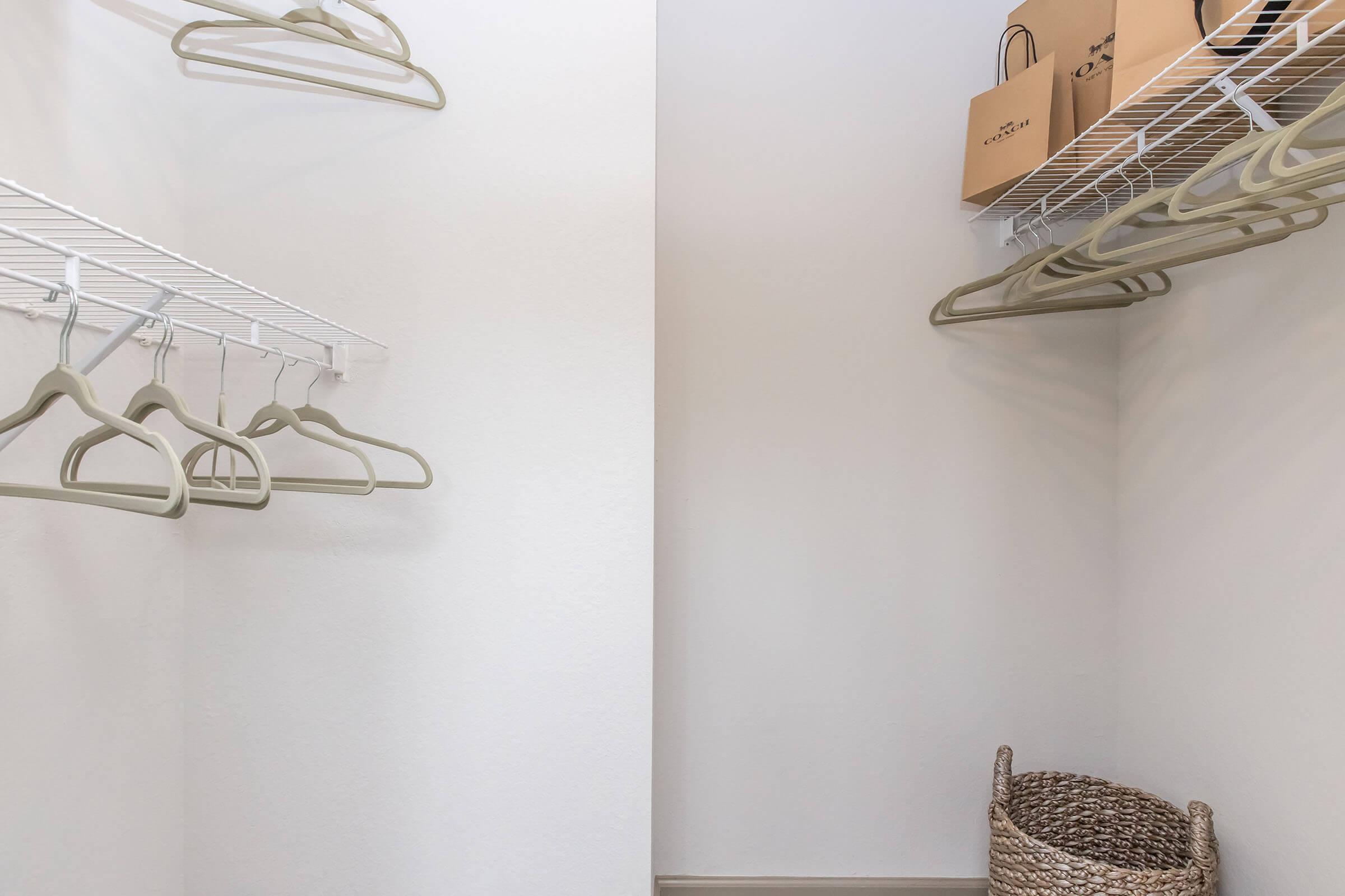 A minimalist closet with white walls and a wire shelving unit. Several light-colored hangers are neatly arranged on the shelving. A decorative woven basket is positioned on the floor, and two boxes sit on the upper shelf, contributing to a tidy and organized appearance.