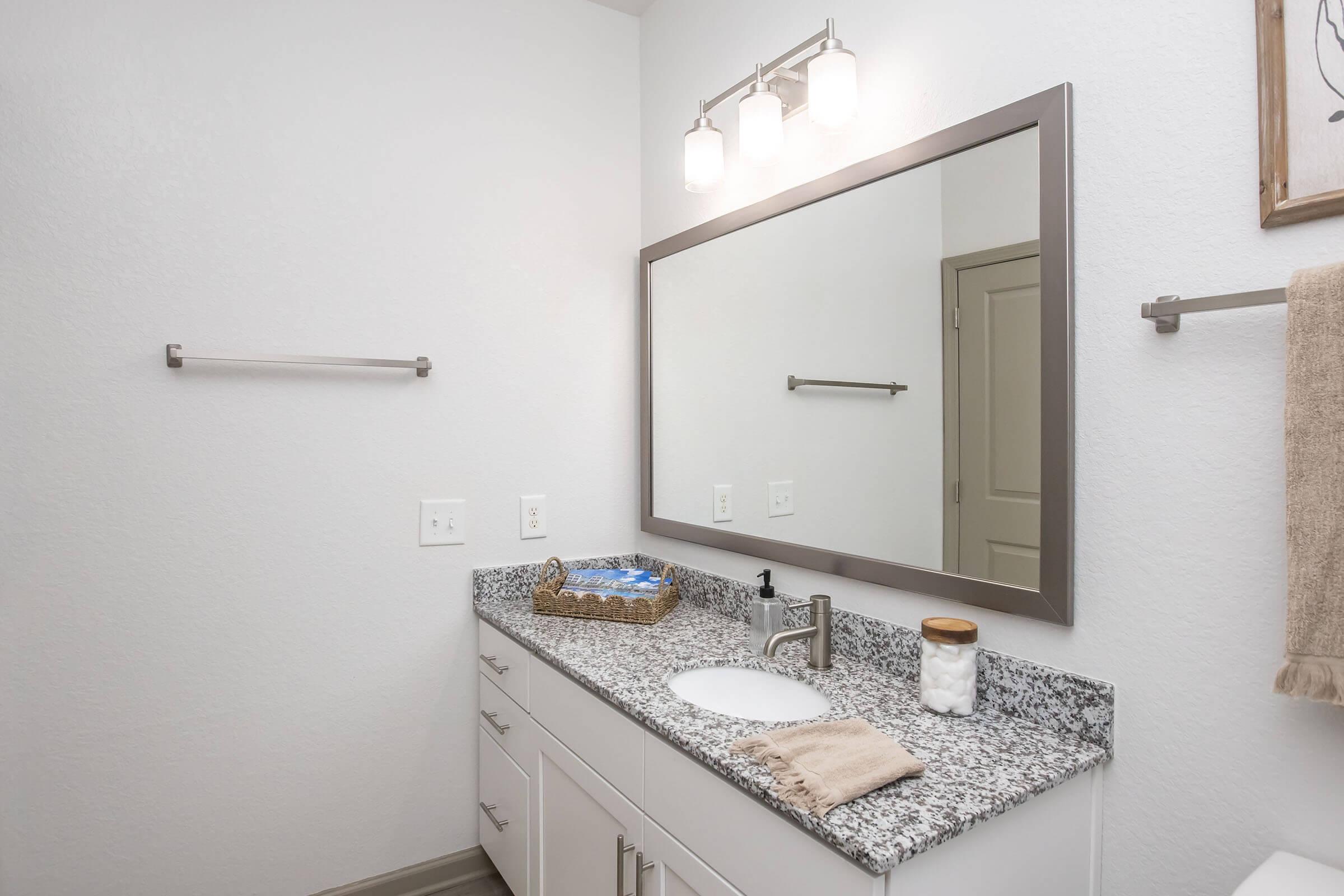 A modern bathroom featuring a granite countertop with a white sink, a large mirror above, and three light fixtures. There is a towel rack on the wall, a decorative basket with towels, and a jar of cotton balls. The walls are painted white, and the floor appears to be a light-colored tile.