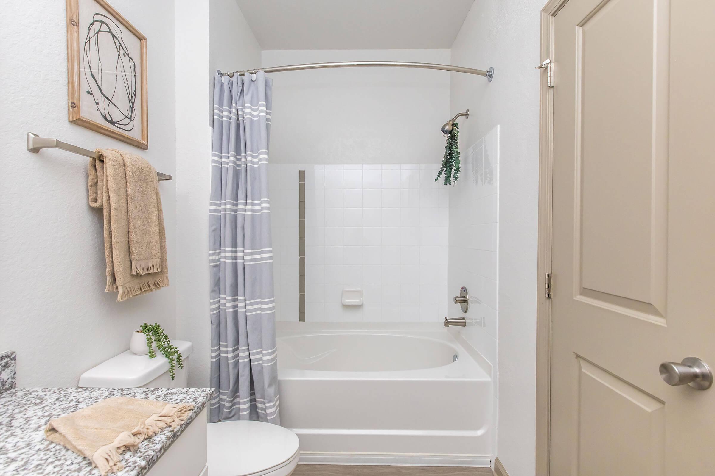 A clean and modern bathroom featuring a white bathtub with a shower curtain, light gray walls, a granite countertop with toiletries, and a towel hanging on a rack. Decor includes a framed wall art piece and a small plant for a refreshing touch.