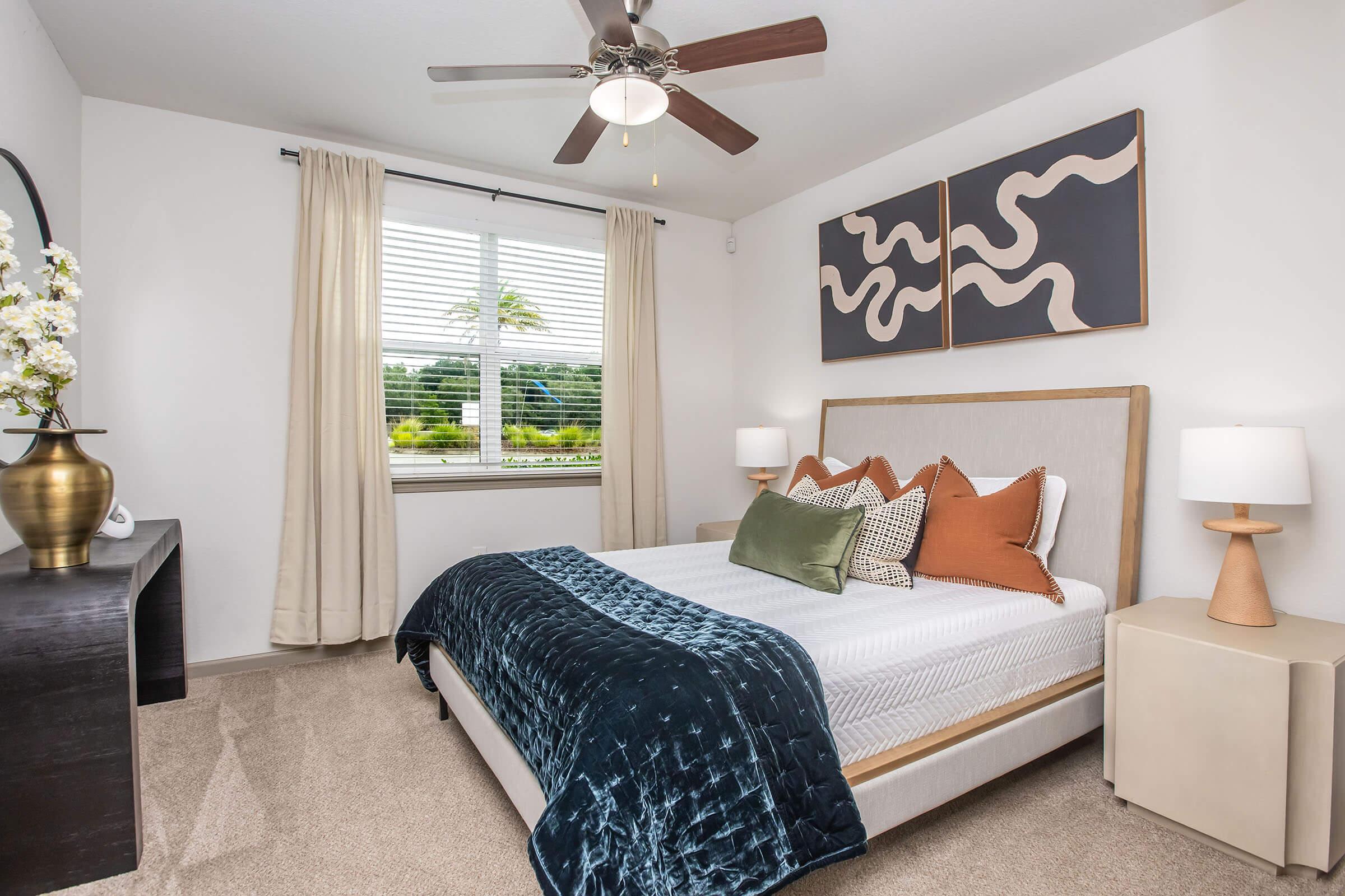 A modern bedroom featuring a bed with a blue throw blanket and decorative pillows in earthy tones. There are two bedside lamps, a dark wooden nightstand, and abstract wall art above the bed. Natural light enters through a window with sheer curtains, which overlooks greenery outside.