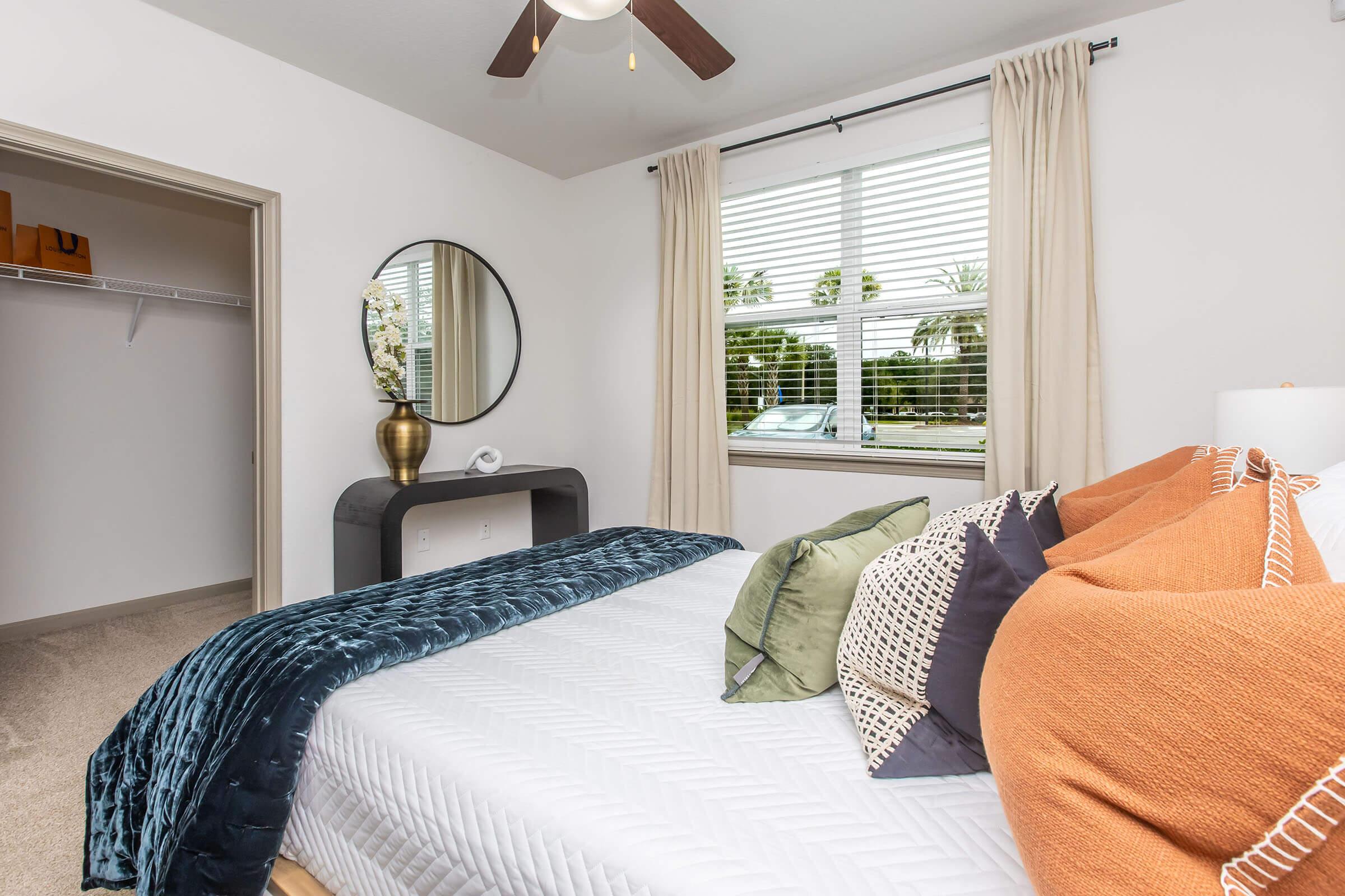 A well-decorated bedroom featuring a queen-sized bed with a white quilt and colorful pillows. There's a round mirror above a sleek black nightstand, a large window with sheer curtains allowing natural light, and a closet with open shelving visible in the background. The overall decor is cozy and inviting.