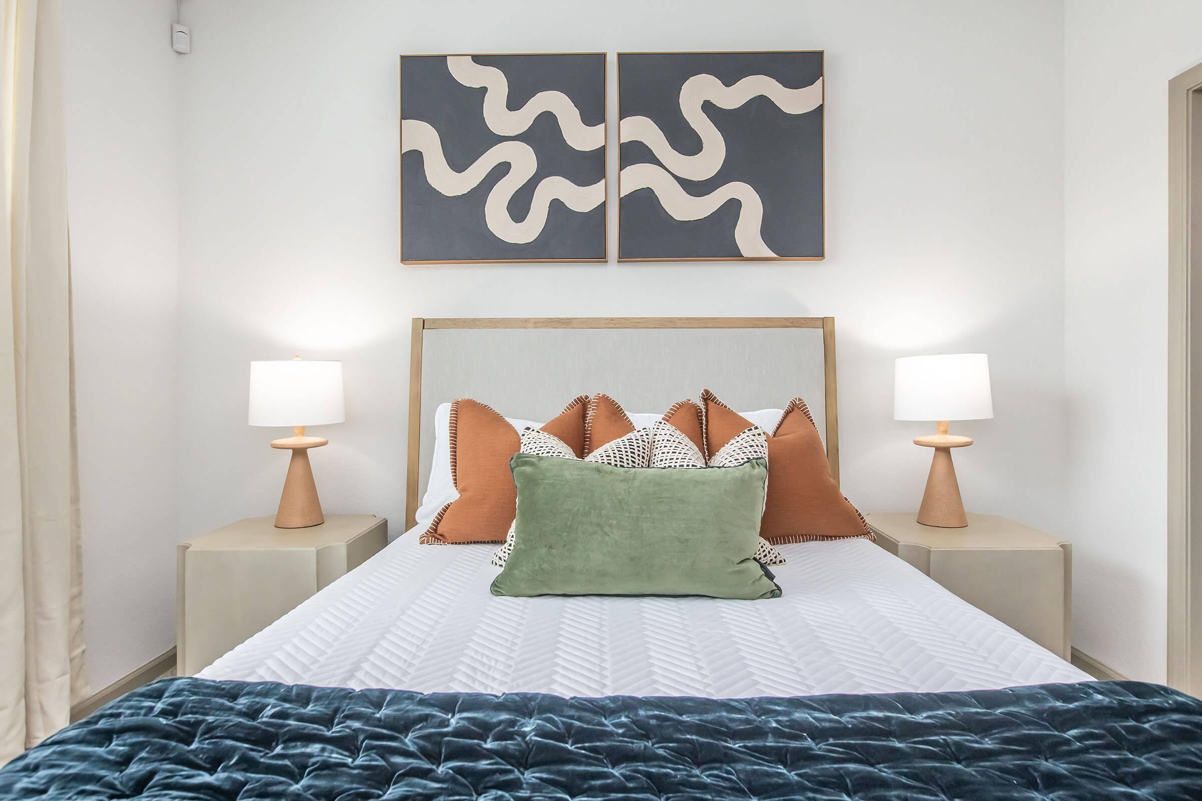 A stylish bedroom featuring a bed with white bedding adorned with colorful throw pillows. On either side of the bed are modern bedside lamps with wooden bases. Above the bed, abstract artwork in dark tones with wavy designs adds a contemporary touch. Soft lighting creates a cozy atmosphere.