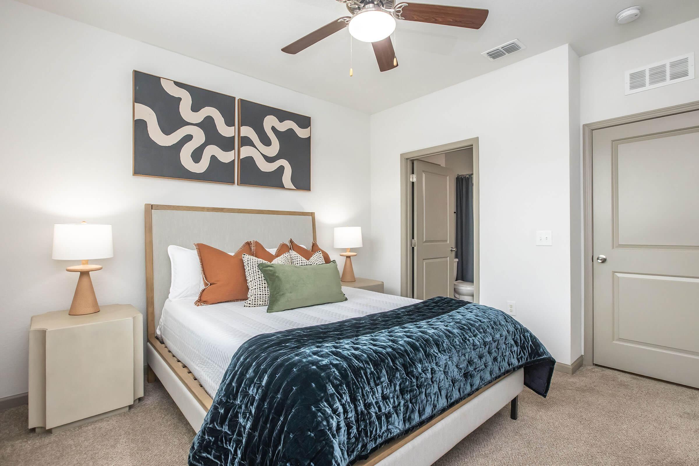 A modern bedroom featuring a neatly made bed with a dark blue velvet blanket and decorative pillows in shades of green and orange. There are two lamps on bedside tables and abstract wall art above the headboard. A door leads to a bathroom, and the walls are painted in neutral tones.