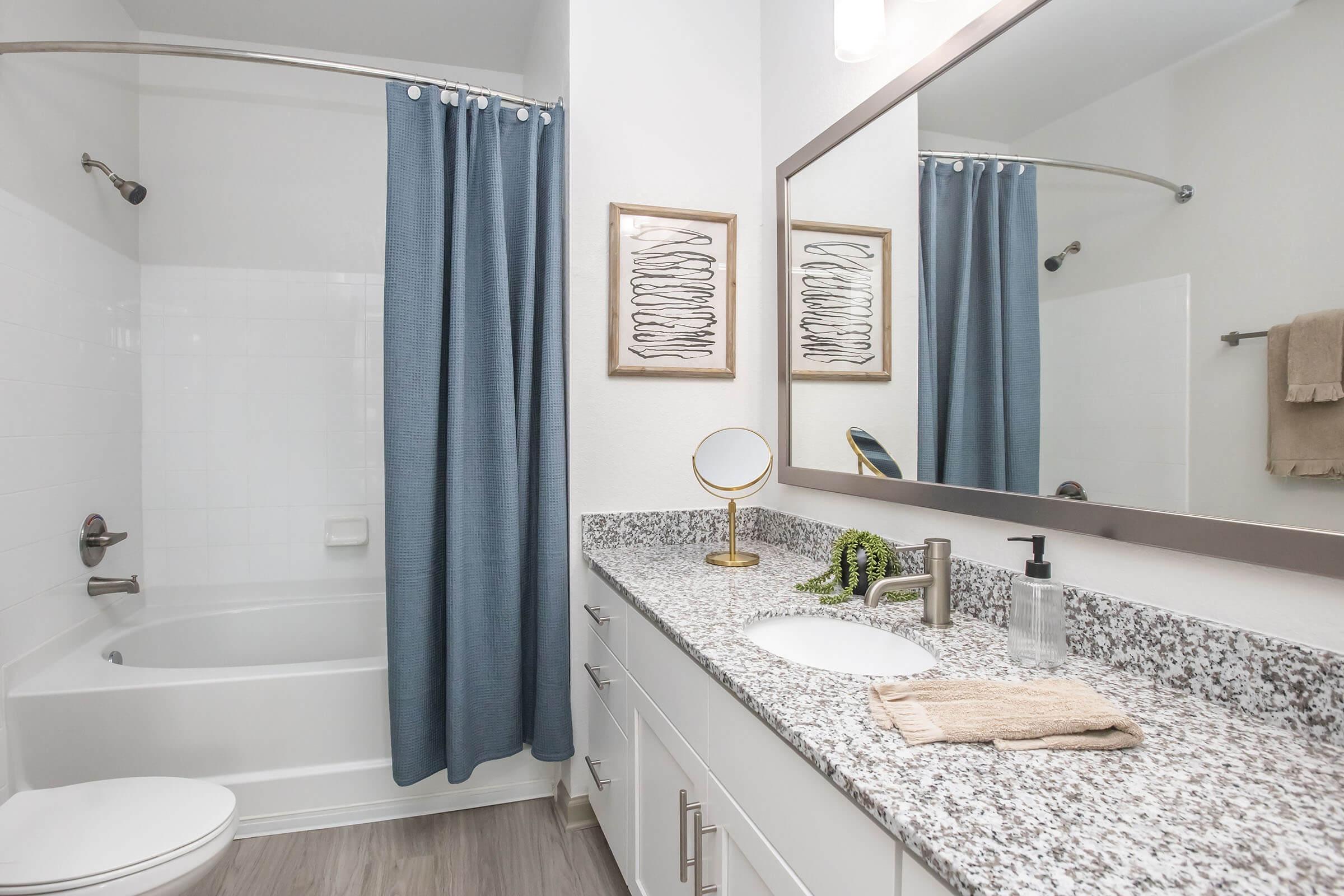 A modern bathroom with a granite countertop, a white sink, and a large mirror. The space includes a shower/tub combo with a blue curtain, a small round mirror, and decorative wall art. There are neatly arranged toiletries and a beige towel hanging on the wall, creating a clean and inviting atmosphere.