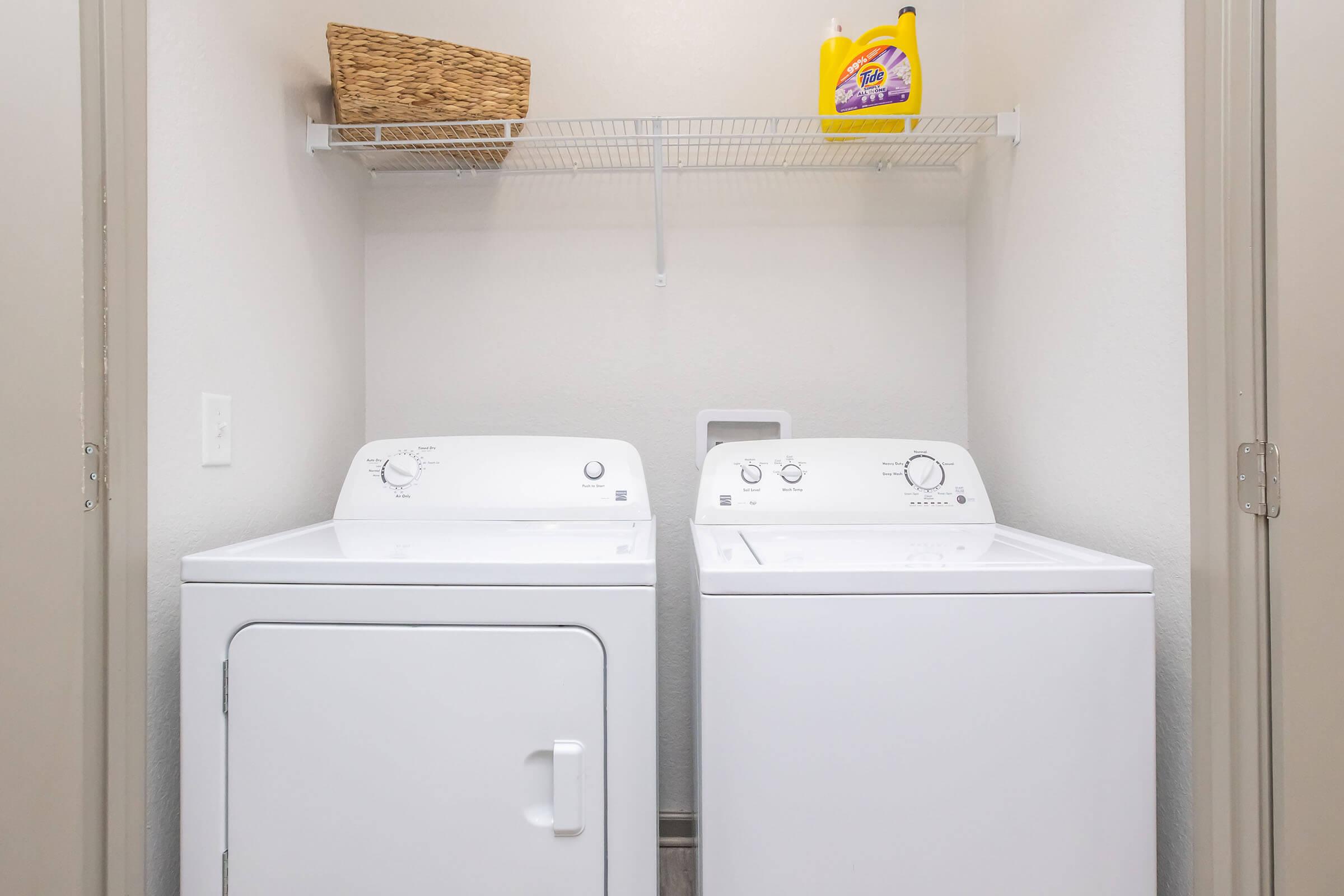 A laundry room featuring a white washing machine and a white dryer side by side. Above them, there is a wall shelf with a woven basket and a bottle of laundry detergent. The walls are painted in a light color, creating a clean and organized appearance.