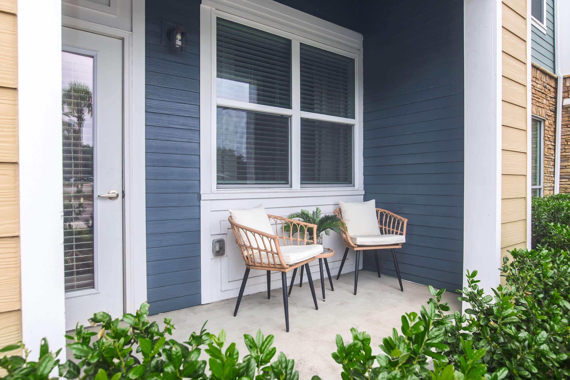 A cozy porch featuring two wicker chairs with cream cushions, a small potted plant between them, and a backdrop of blue and beige house siding. The entrance includes a glass-panel door and a window with white blinds, surrounded by neatly trimmed green shrubs.