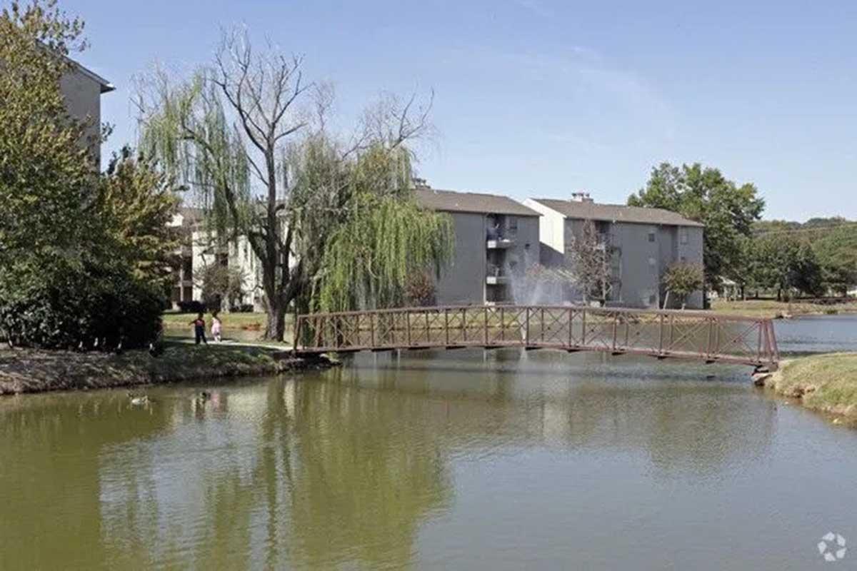 A serene view of a pond surrounded by apartments. A small metal bridge crosses the water, with trees and shrubs lining the shore. People can be seen walking nearby, enjoying the natural surroundings on a clear day with blue skies.