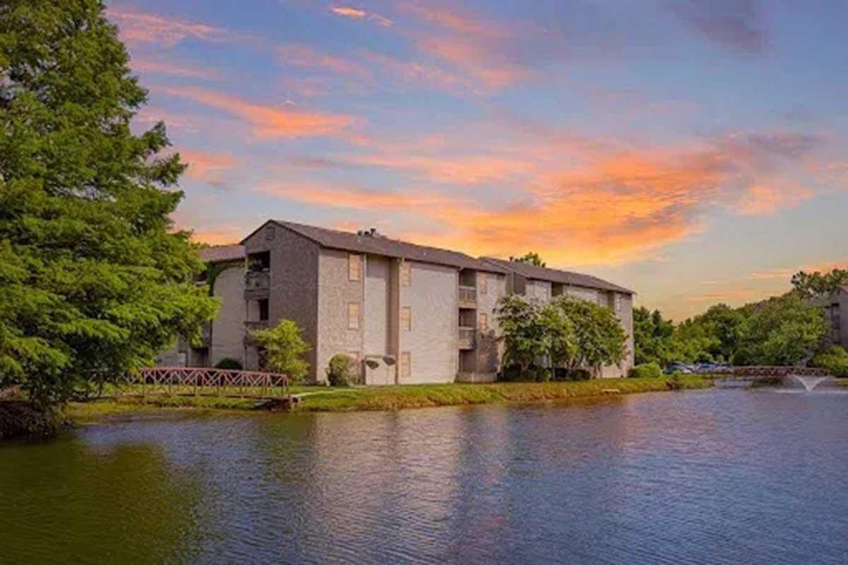 A picturesque view of a modern apartment building beside a serene body of water, surrounded by lush greenery. The sky is ablaze with vibrant orange and purple hues during sunset, creating a tranquil and inviting atmosphere.