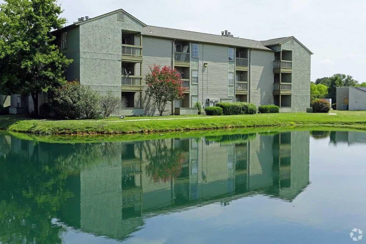 A three-story gray apartment building with balconies overlooks a calm, reflective pond surrounded by lush green grass and trees. The sky is partly cloudy, creating a serene residential scene.