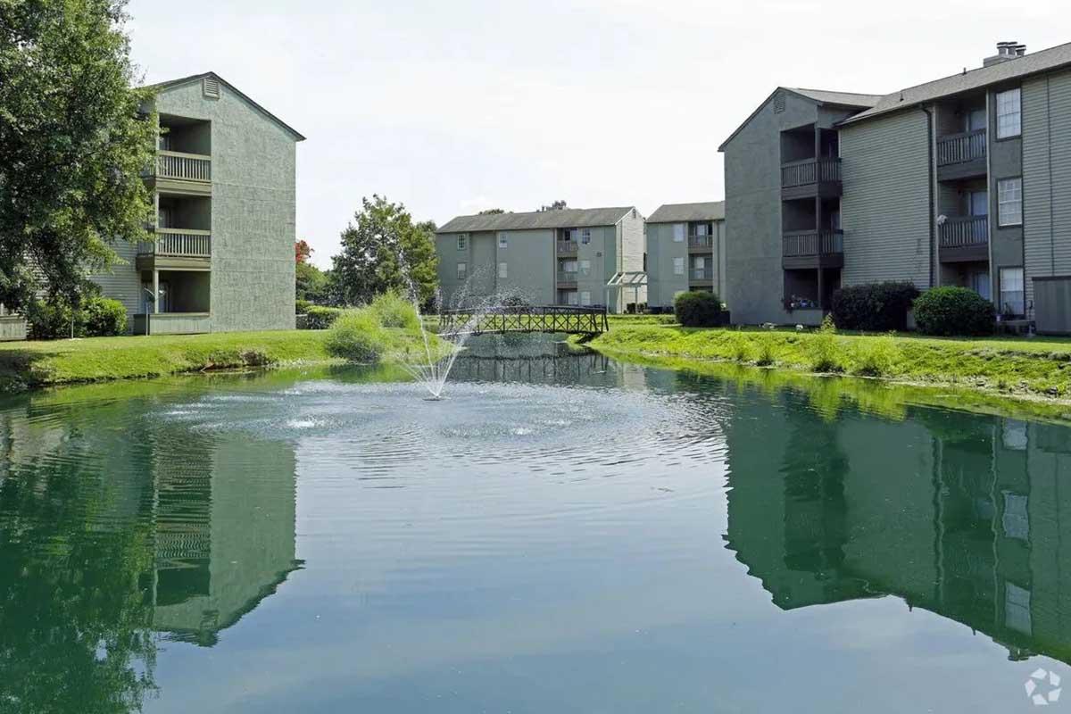A serene pond surrounded by greenery and apartment buildings. The pond features a small fountain creating ripples on the water's surface. Lush grass borders the water, and a wooden bridge connects the two sides, enhancing the tranquil atmosphere of the community.