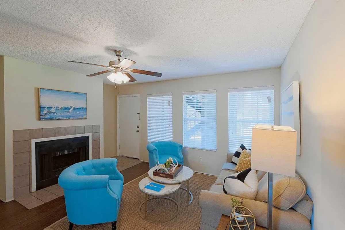A cozy living room featuring a light-colored wall, two bright blue armchairs, a beige sofa, a round coffee table, and a lamp. The space has a ceiling fan and large windows with white blinds. A fireplace is set against a tiled wall, and decorative items are arranged on the coffee table.