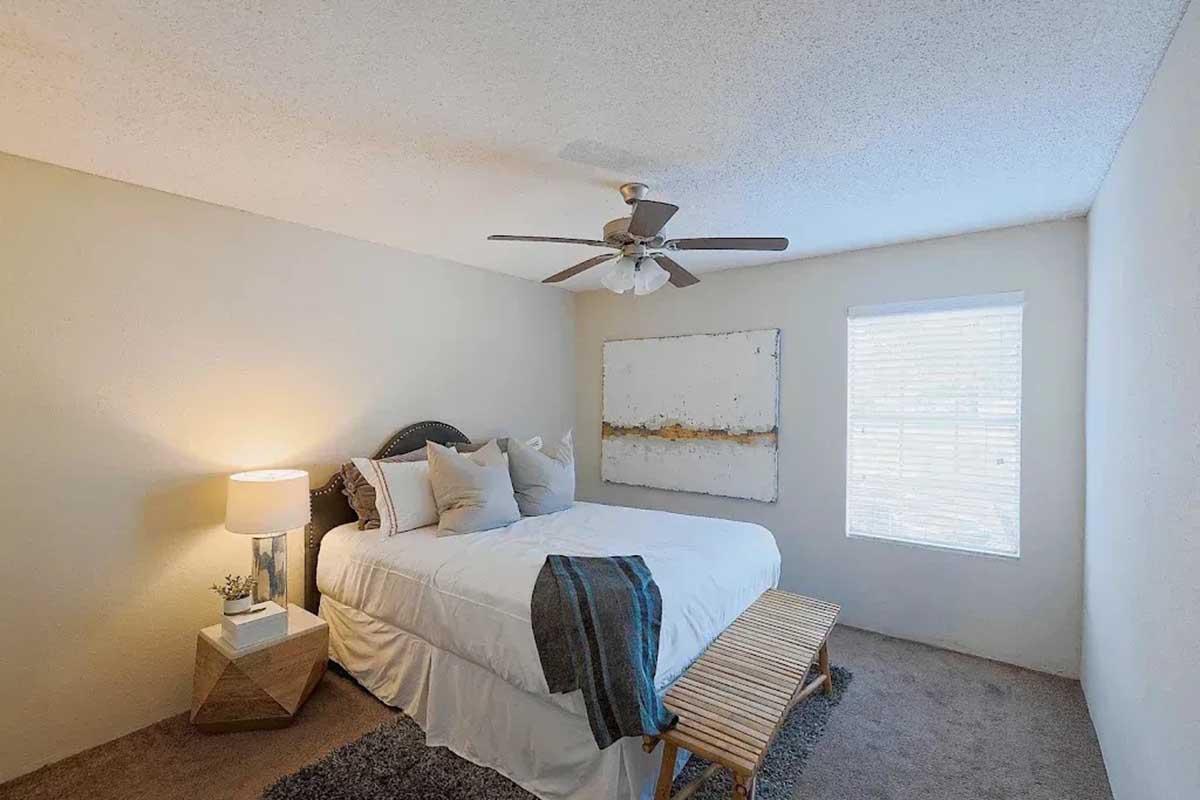 Cozy bedroom featuring a queen-size bed with decorative pillows, a small bedside table with a lamp, a wooden bench at the foot of the bed, and a window with blinds. The walls are a soft beige, and there is a ceiling fan above. A large abstract painting adorns one wall, adding a touch of decor.