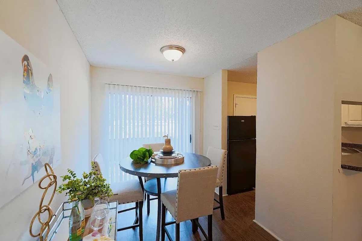 A small dining area featuring a round table with four upholstered chairs, a decorative centerpiece, and a nearby plant. There's a window with vertical blinds allowing natural light, and a black refrigerator can be seen in the background, along with light-colored walls and wooden flooring.