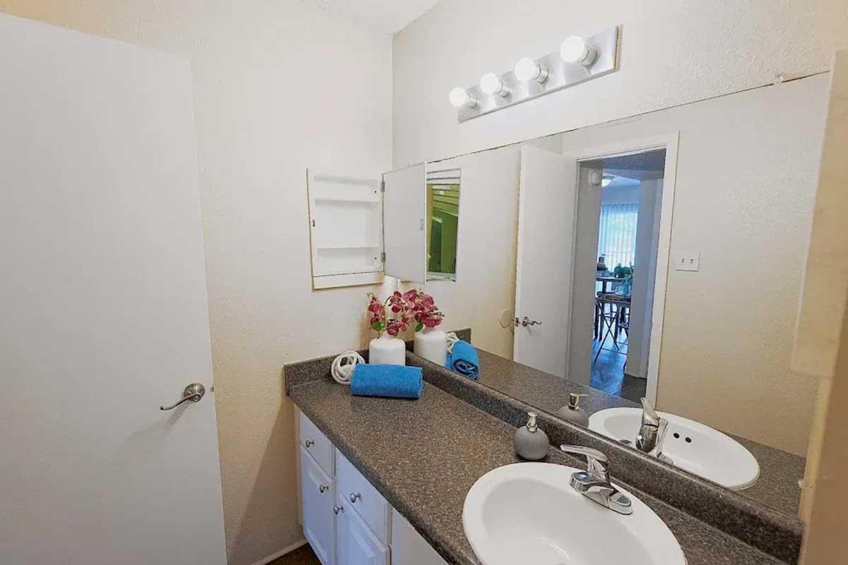 A clean, bright bathroom featuring a dark countertop, white cabinetry, and a sink. There is a mirror above the sink and a small shelf on the wall. A blue towel and several decorative items, including a vase with flowers, are neatly arranged. Natural light is coming from a nearby window.