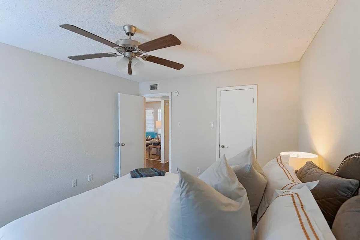 A cozy bedroom featuring a large bed with decorative pillows, a ceiling fan, and warm lighting from a bedside lamp. There are two doors leading to other rooms, and a neutral color palette creates a calm atmosphere. A glimpse of another room is visible through the open door.