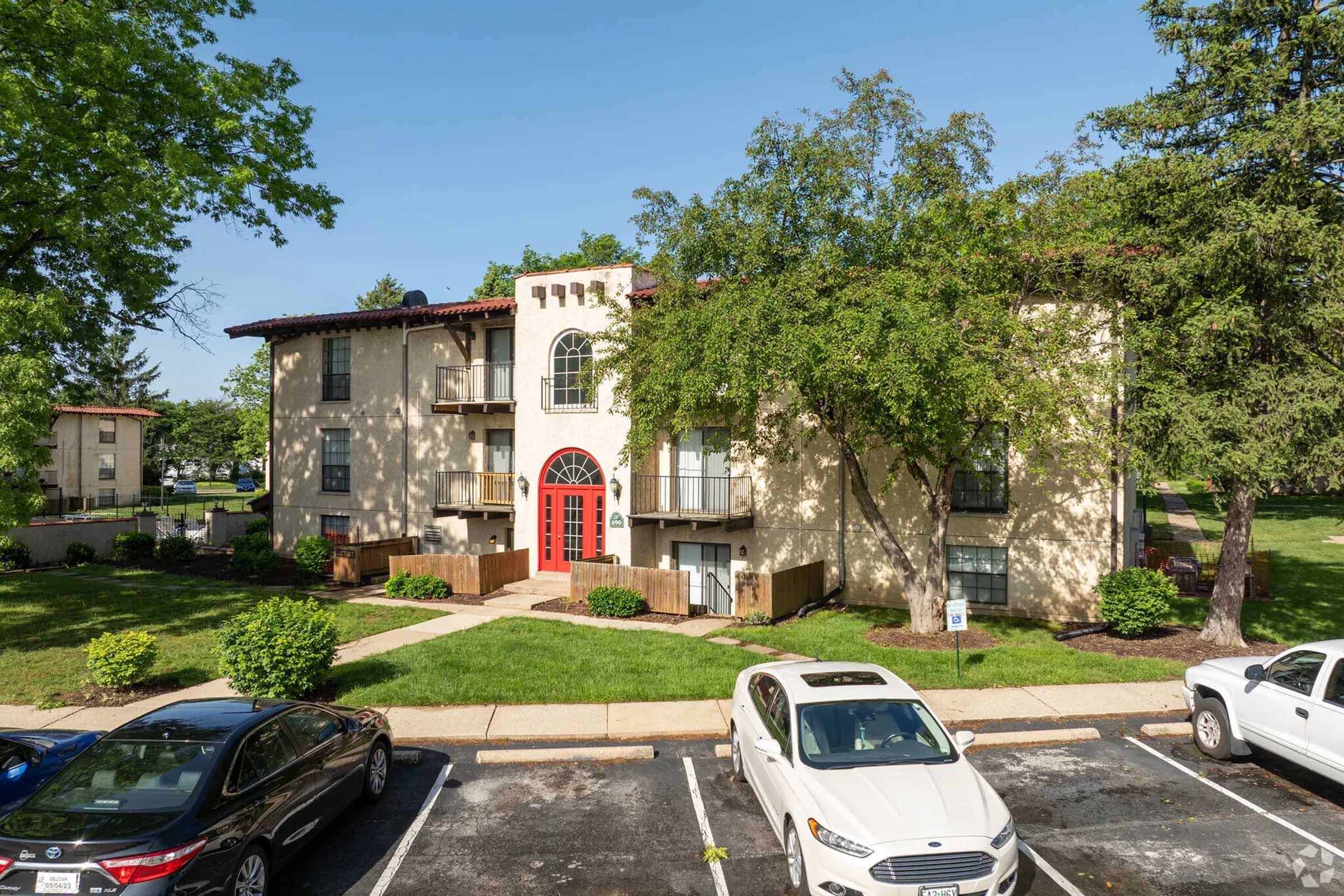 A modern apartment building with a cream-colored facade, red arched entrance, and balconies. Large windows and green trees surround the building, set in a landscaped area with grassy lawns. Two parked cars are visible in the foreground.