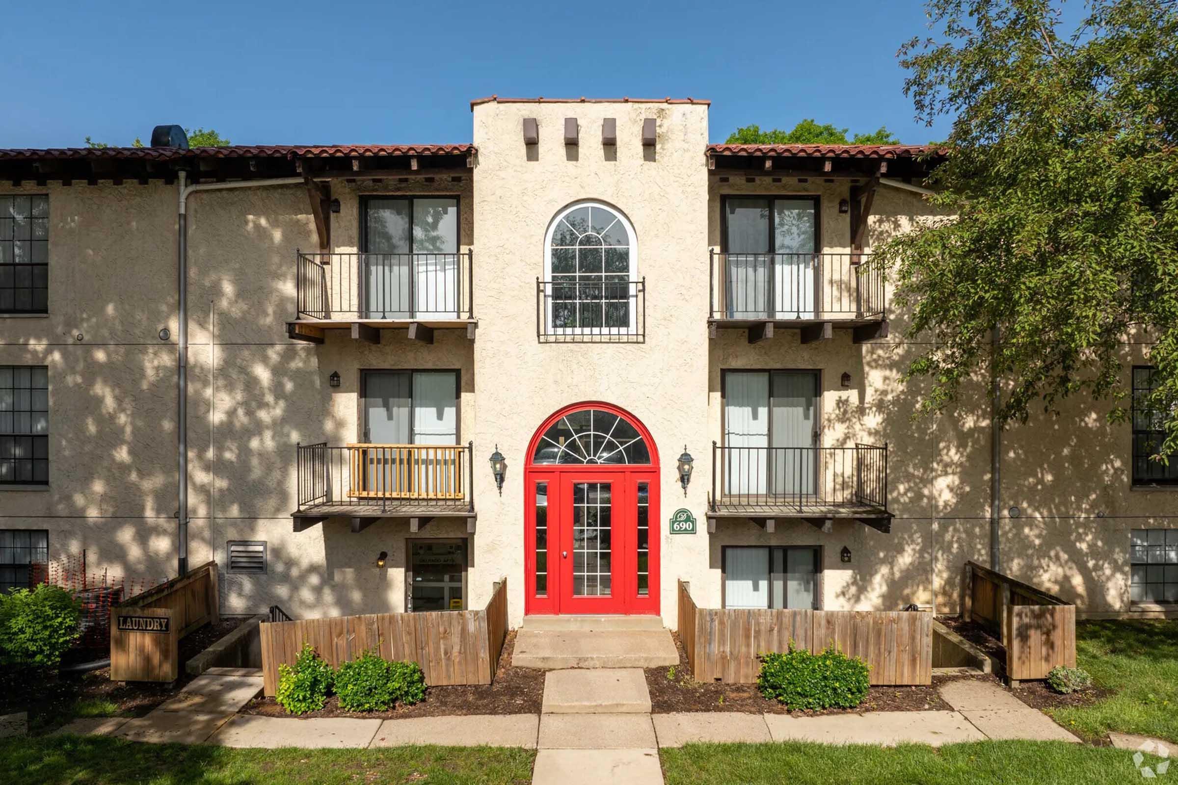 A two-story apartment building with a textured beige exterior and red accents. The entrance features a bright red door framed by arched windows, flanked by small balconies. Lush greenery surrounds the entrance, with wooden fencing and a paved walkway leading up to the door. Clear blue sky overhead.