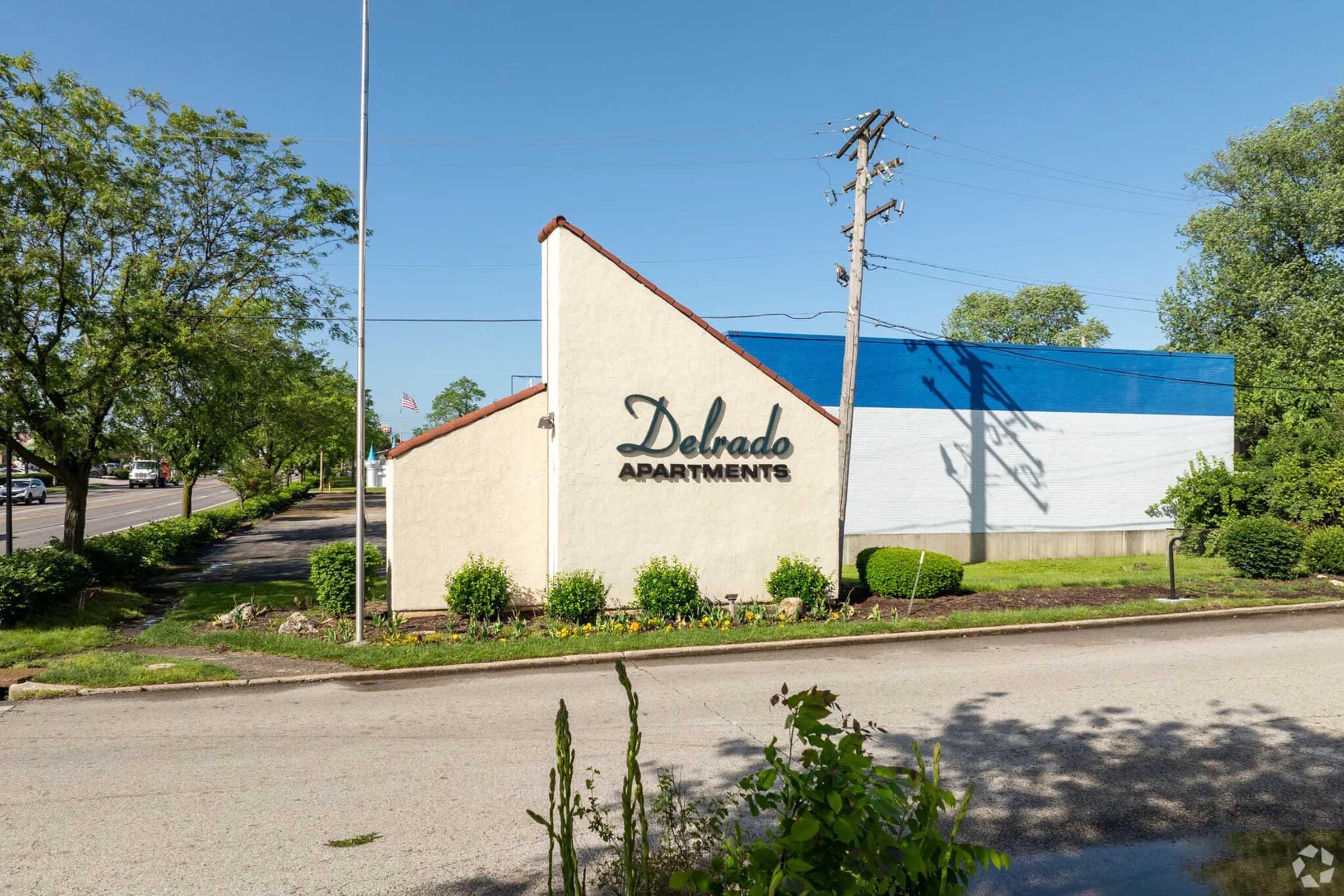 A building sign for "Delrado Apartments" against a clear blue sky, with landscaped greenery in front. The structure features a white wall with blue accents, and utility poles visible nearby. The setting suggests an apartment complex located along a road.