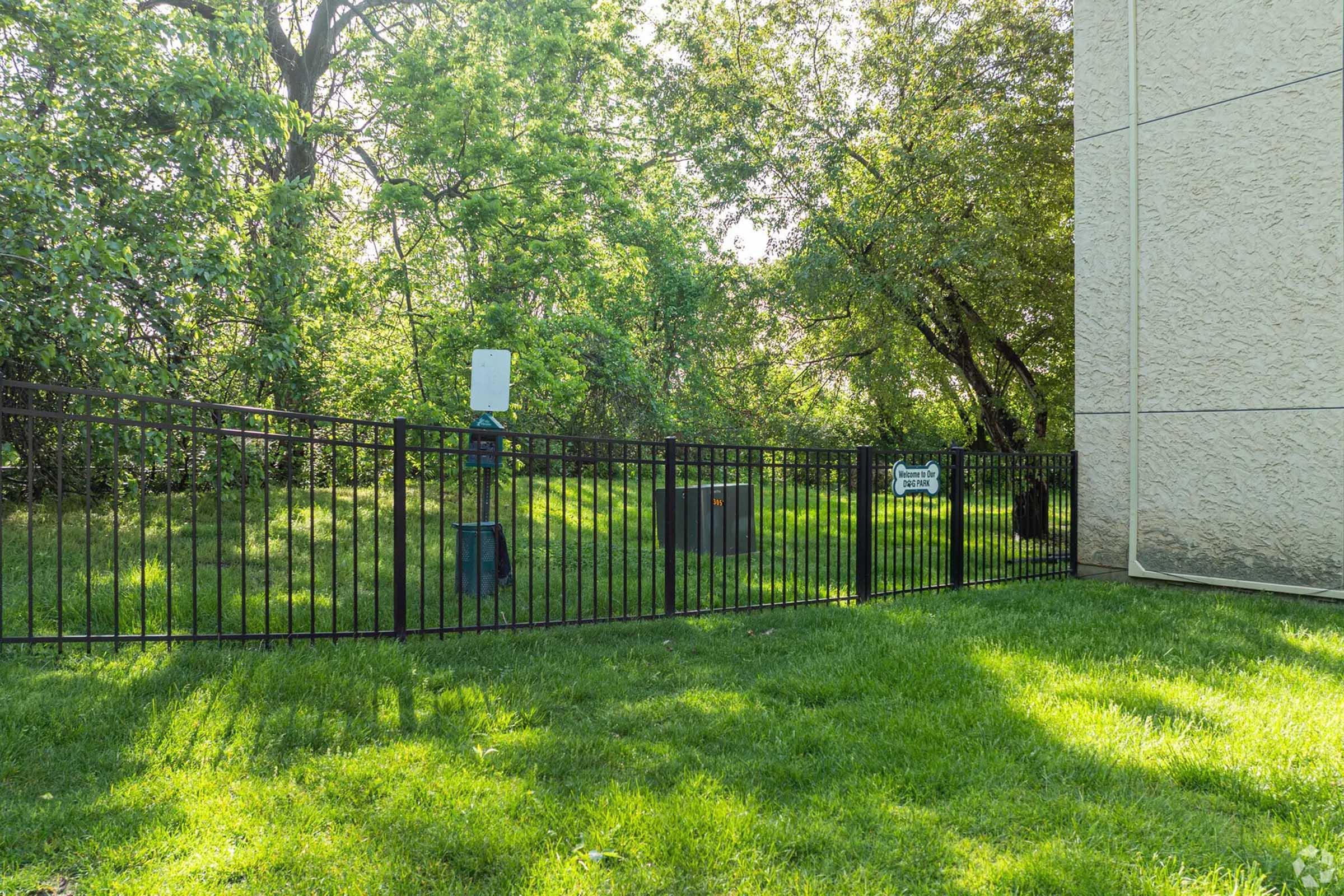 A fenced area beside a building, featuring a grassy lawn and trees in the background. There are two boxes inside the fence, one with a sign, and a small device mounted on a post. The scene is bright and sunny, surrounded by lush greenery.