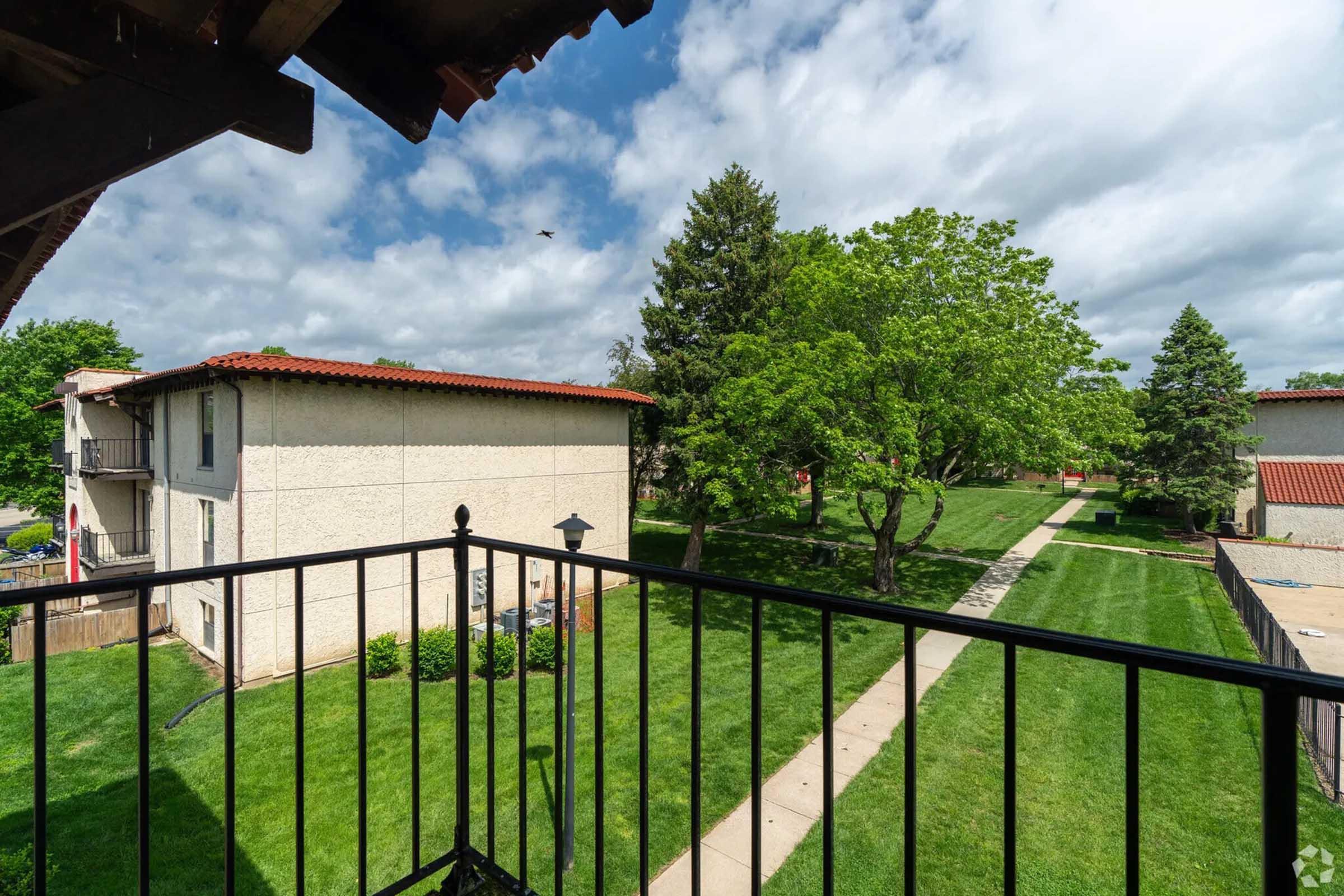 View from a balcony overlooking a green landscape with trees and a sidewalk. Two multi-unit buildings are visible, one with a red roof, and the sky is partly cloudy. The scene conveys a peaceful suburban environment.