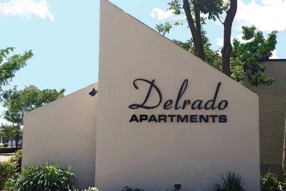 Sign for Delrado Apartments, featuring curved architecture and surrounded by greenery. The sign is prominently displayed, showcasing the name "Delrado" in a stylish font with the word "APARTMENTS" below it. Bright blue skies and a few clouds are visible in the background.