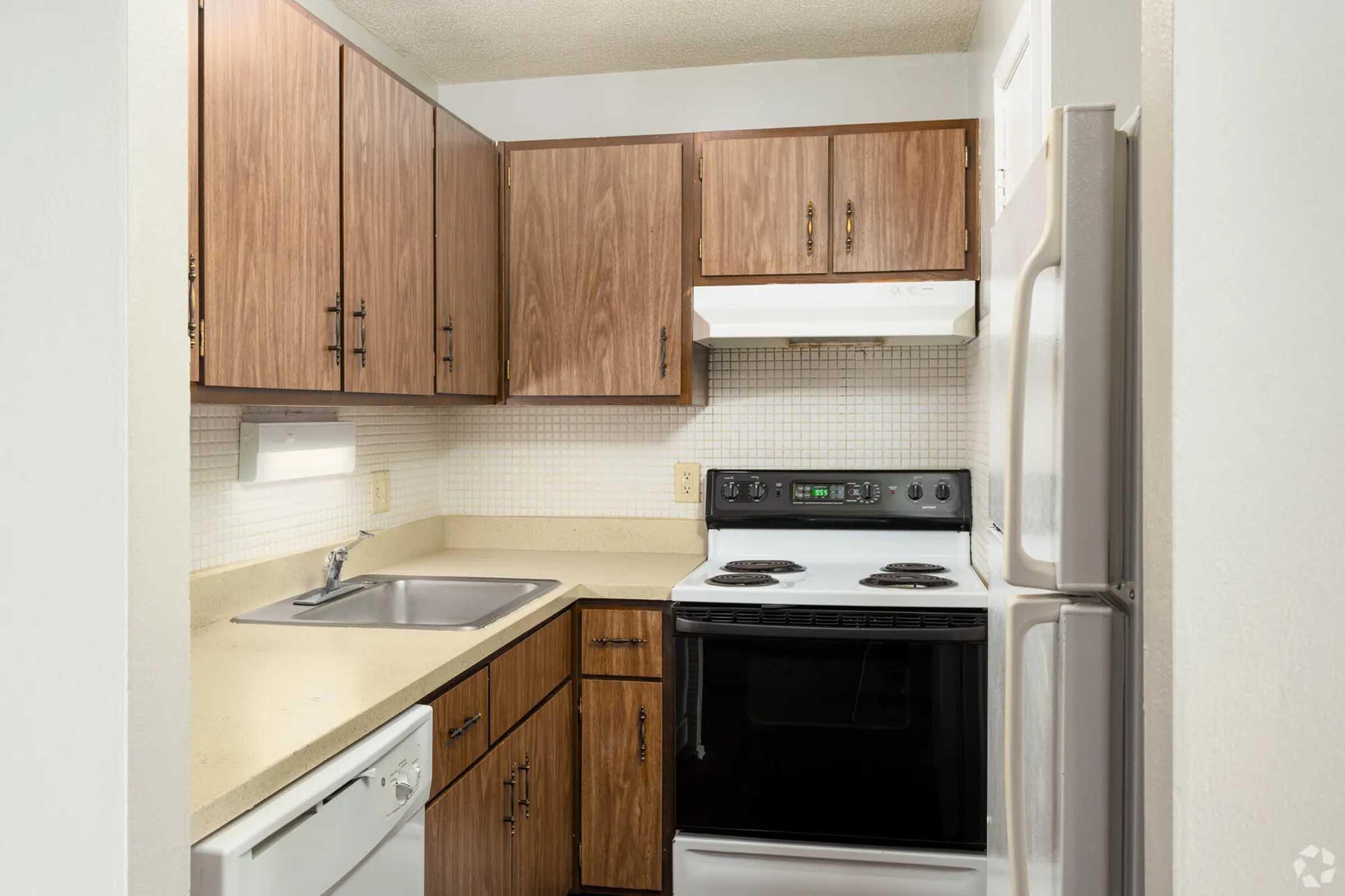 A small kitchen featuring wooden cabinets, a sink, a countertop, and a white tile backsplash. It includes a built-in stove with an oven, a microwave above, and a dishwasher. The overall design is simple and functional, with a focus on efficiency in a compact space.