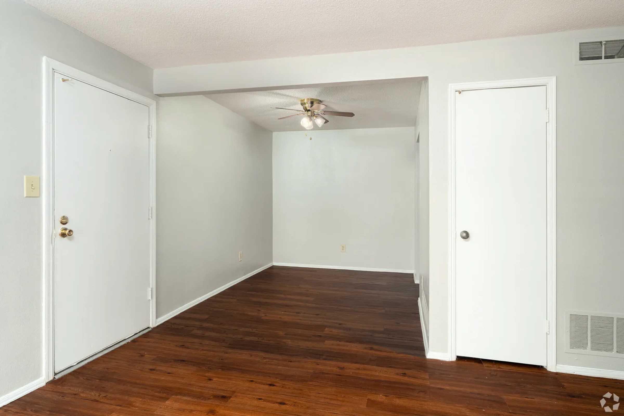 A light-colored room with hardwood flooring, featuring a ceiling fan and two doors on the left side, one leading outside and the other to a closet. The walls are painted in a soft gray, creating a spacious and inviting atmosphere. Natural light enters from an unseen window.