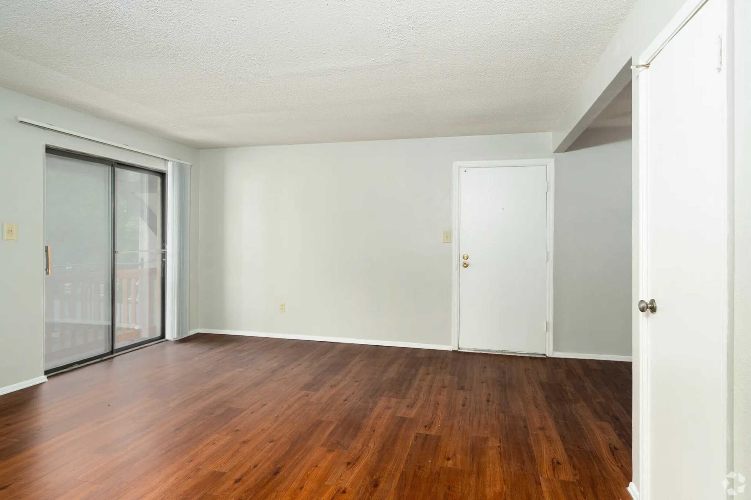 A spacious, empty room with light gray walls and a smooth, wooden floor. There is a large sliding glass door leading to a balcony on one side and a white door on the other side. The ceiling has a textured finish, contributing to a bright and open atmosphere.