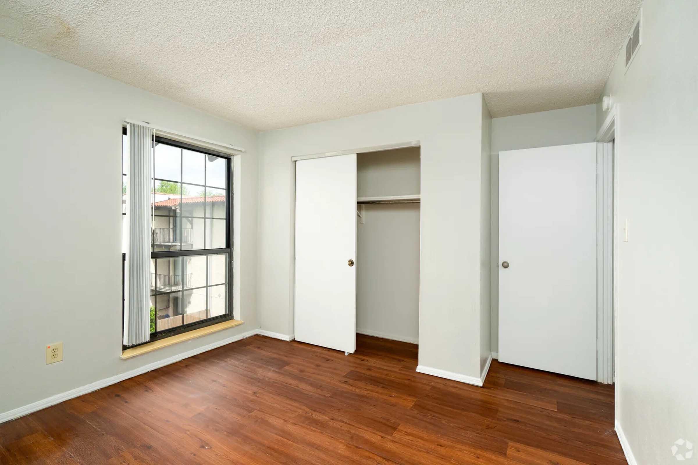 Empty bedroom featuring light-colored walls, a large window with vertical blinds, and laminate flooring. There are two sliding closet doors, one partially open, and a sunny atmosphere. The room has a minimalistic design with neutral tones.