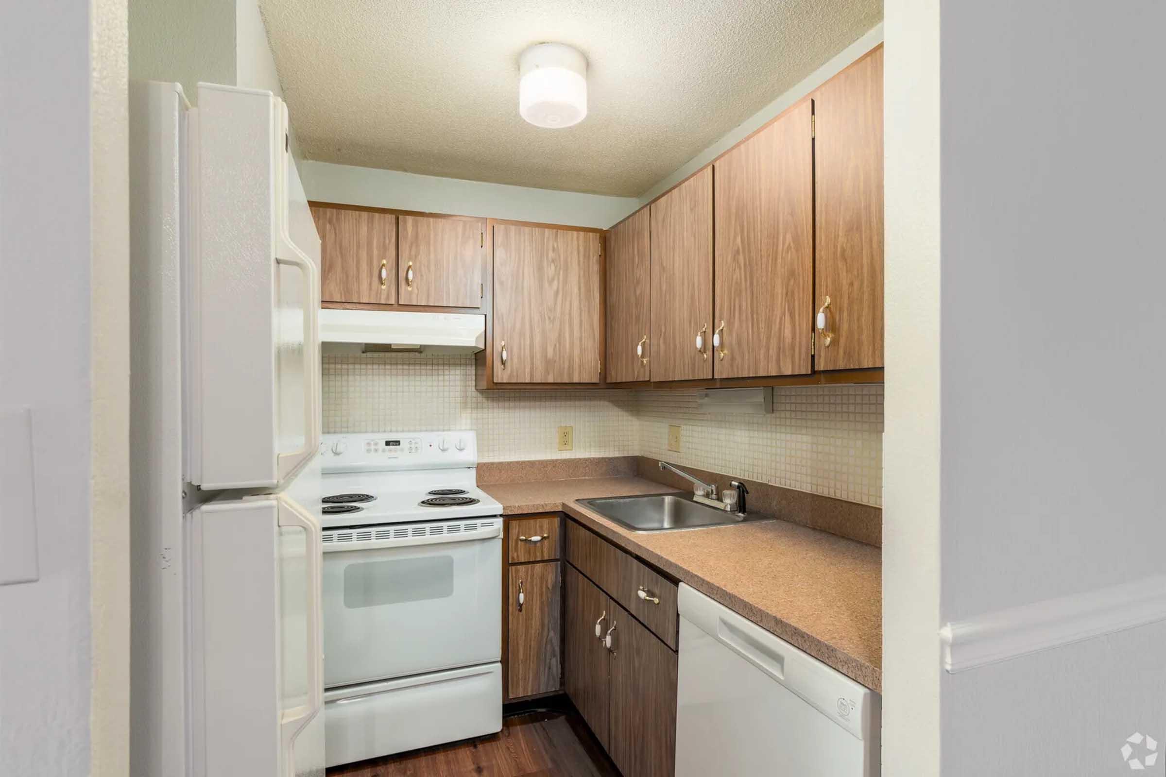 A small, modern kitchen featuring brown wooden cabinets, a white stove and microwave, a stainless steel sink, and a dishwasher. The countertops are light-colored. The floor has a dark wood finish, and there is a ceiling light fixture illuminating the space.